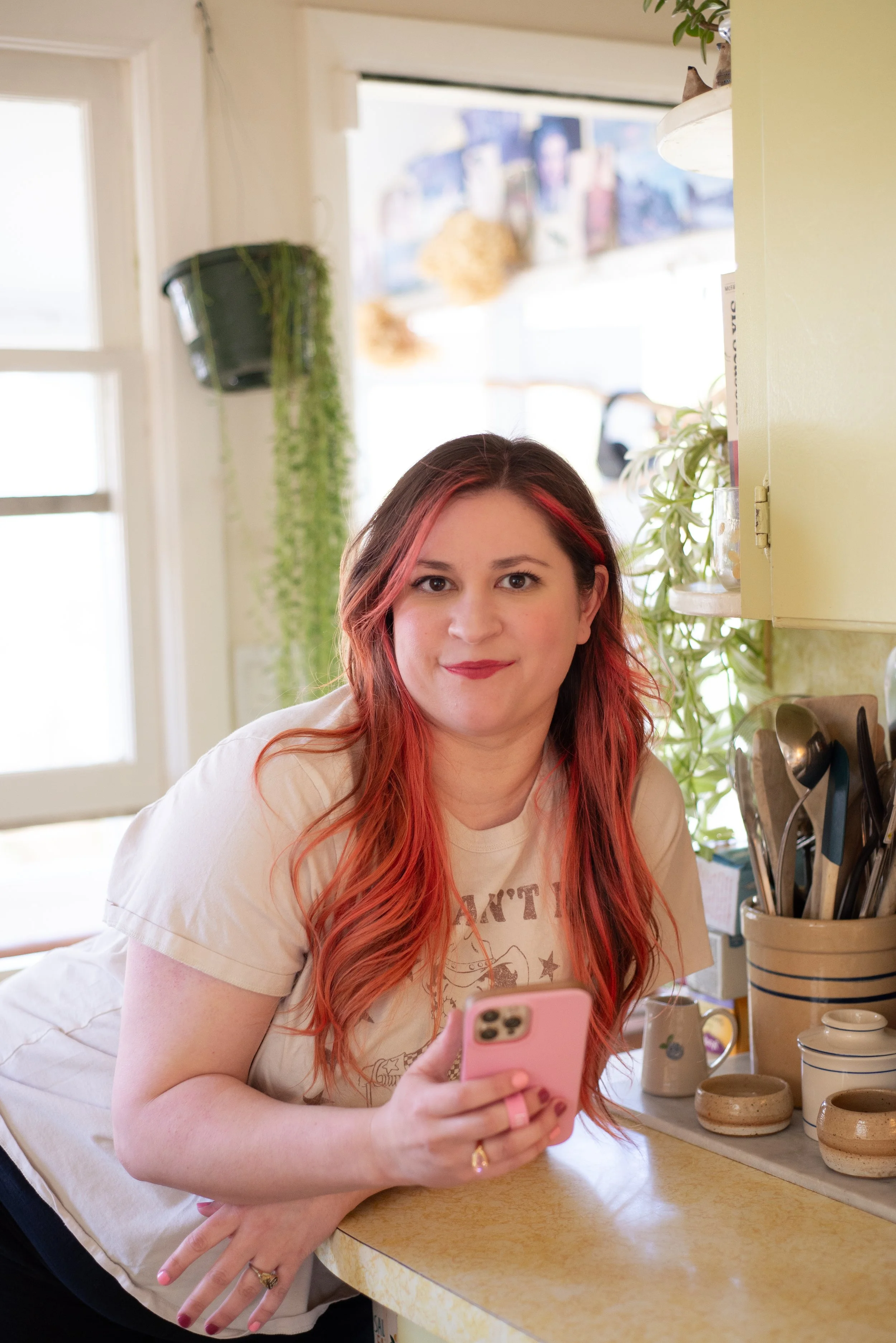 A woman with red hair and a beige t-shirt holding a pink smartphone in a kitchen setting, with cabinets, plants, and kitchen utensils visible in the background.