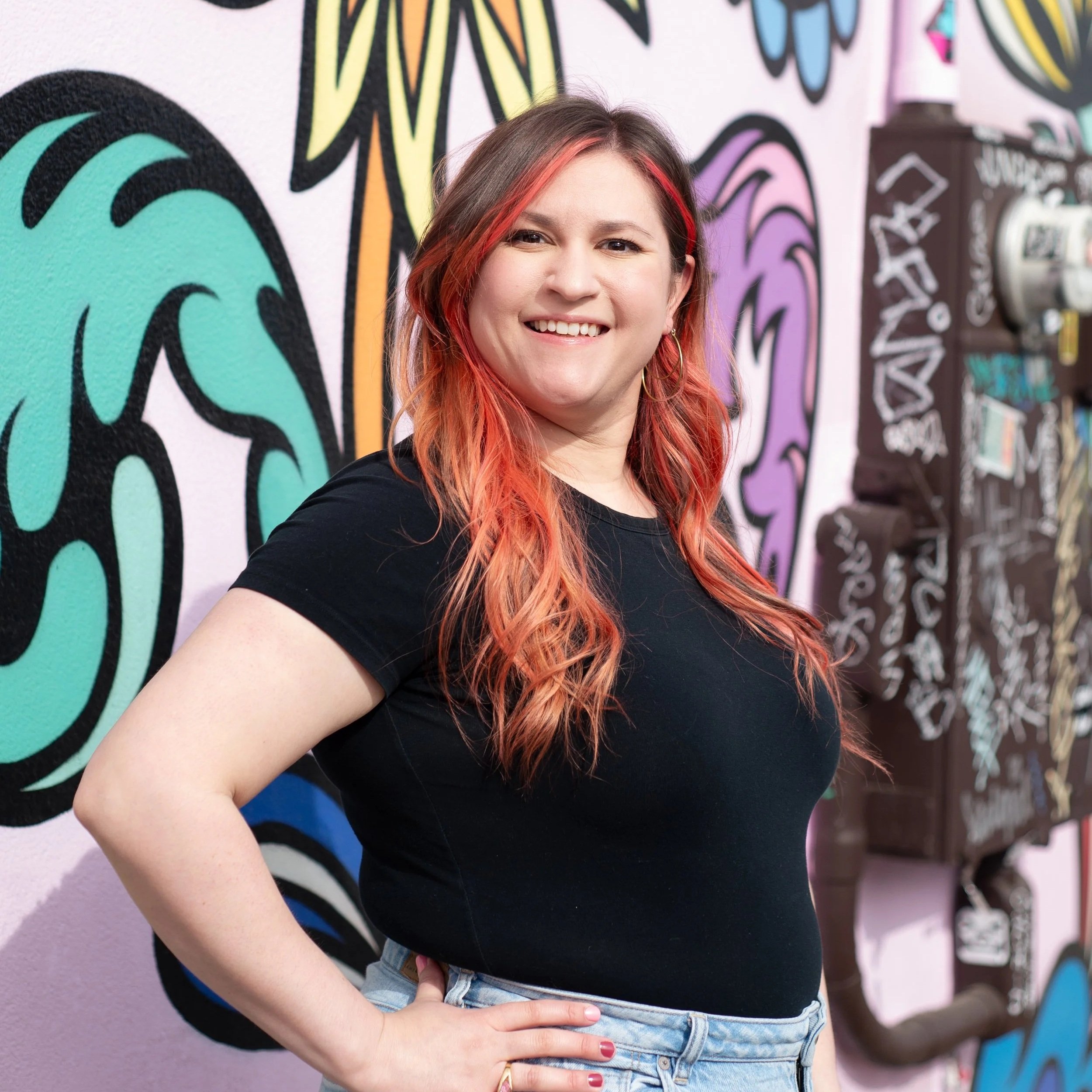 A woman with long red hair, smiling and posing with her hand on her hip, standing in front of a colorful graffiti wall.