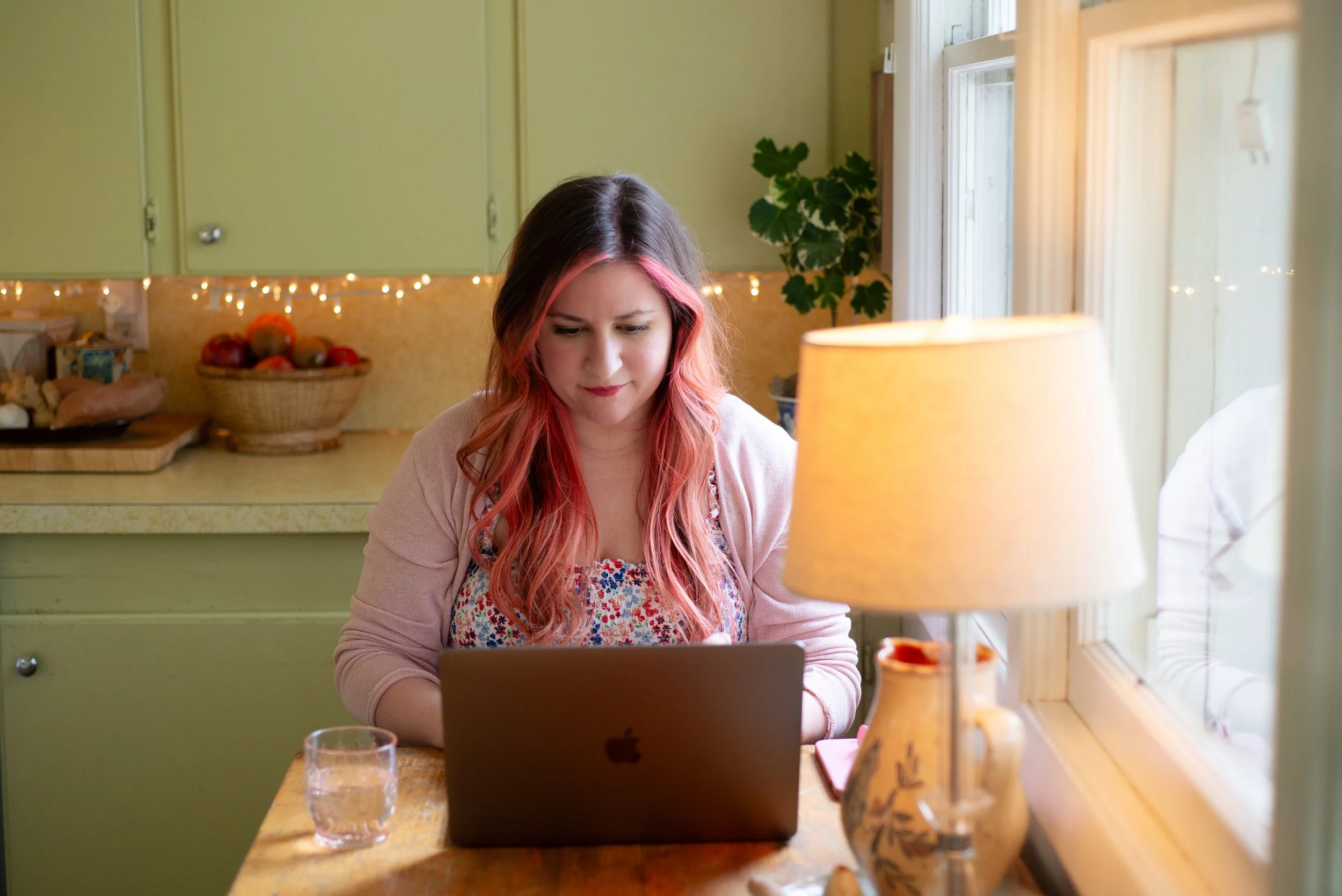A woman with wavy pink hair working on a laptop at a kitchen table near a window, with a glass of water, a lamp, and decorative vases.