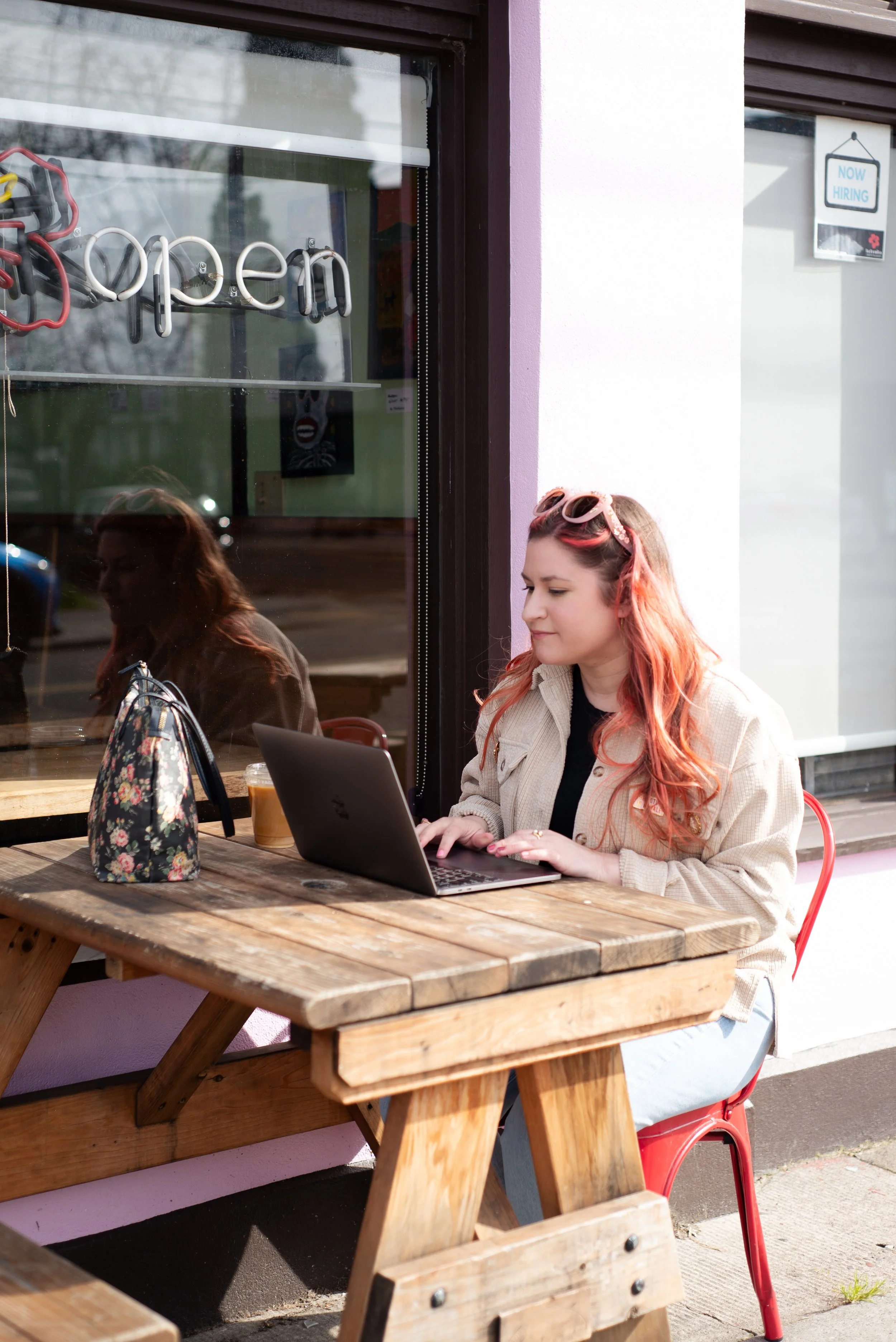 A young woman with pink hair and sunglasses on her head sitting outside at a wooden table, working on her laptop, with a floral bag and iced coffee nearby, outside a cafe with a 'Now Hiring' sign in the window.