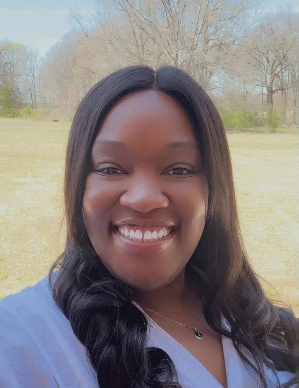 A woman smiling outdoors in a park with trees and grass in the background.