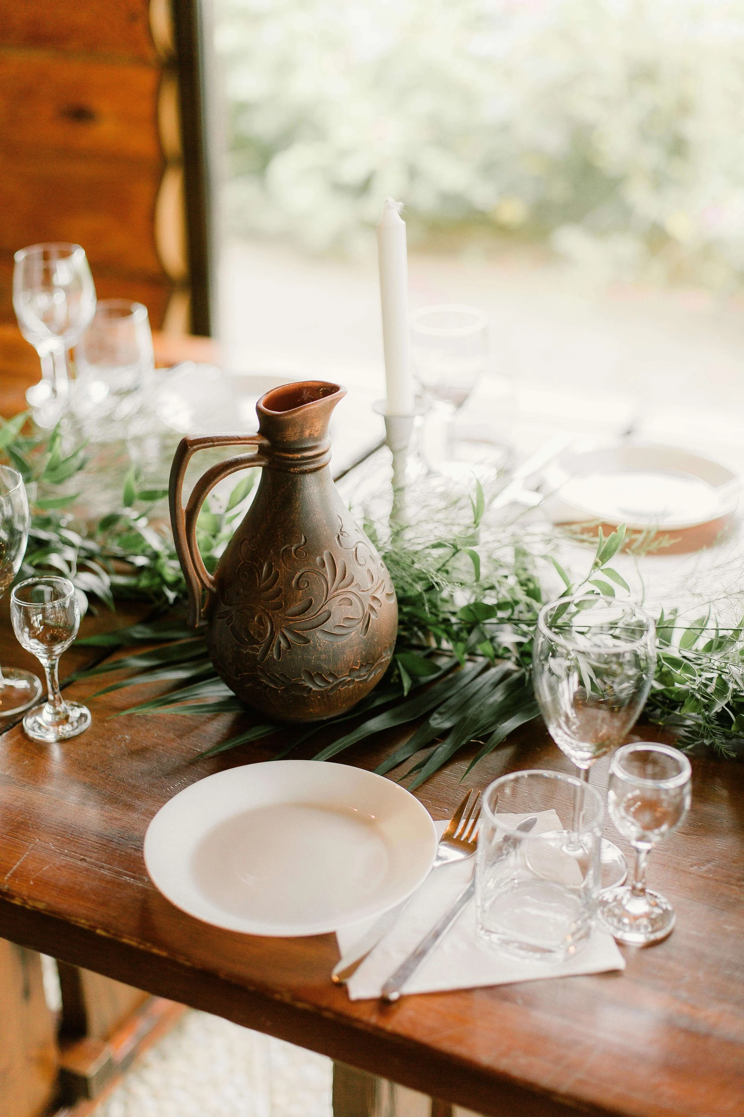 A wooden dining table decorated with a rustic brown vase, green foliage, a single white candle, and multiple empty glasses, set for a meal.