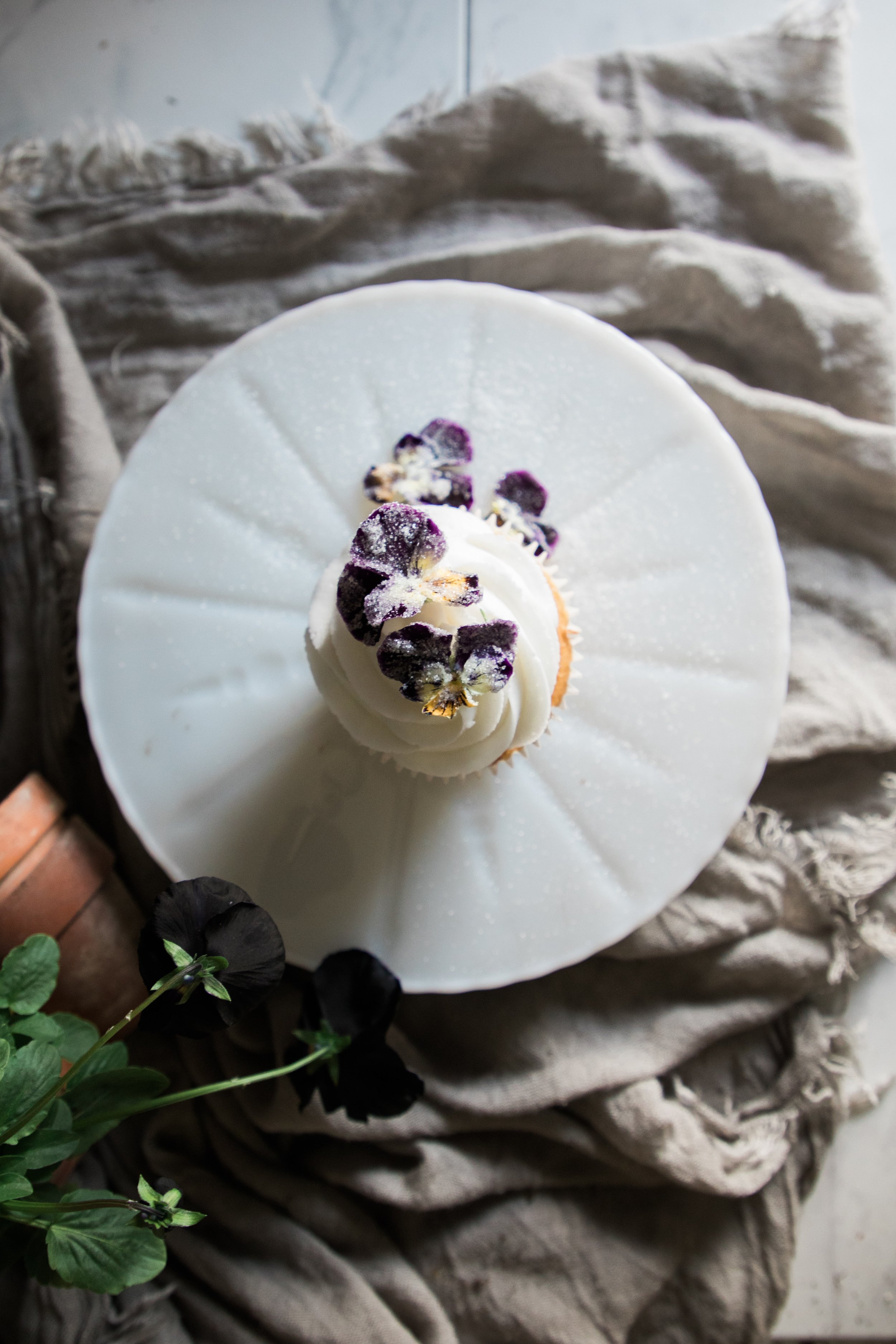 Top-down view of a cupcake topped with white frosting and purple edible flowers, placed on a white cake stand with a speckled surface, surrounded by a beige cloth and a small green potted plant.