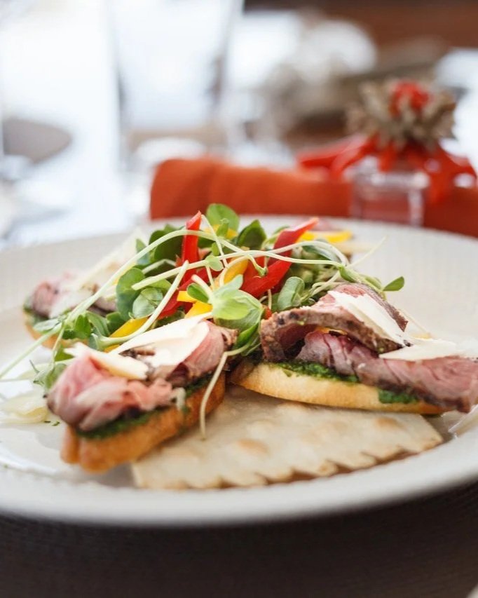 Steak served on a plate with greens and colorful chopped peppers, with a red napkin and glass in the blurry background.
