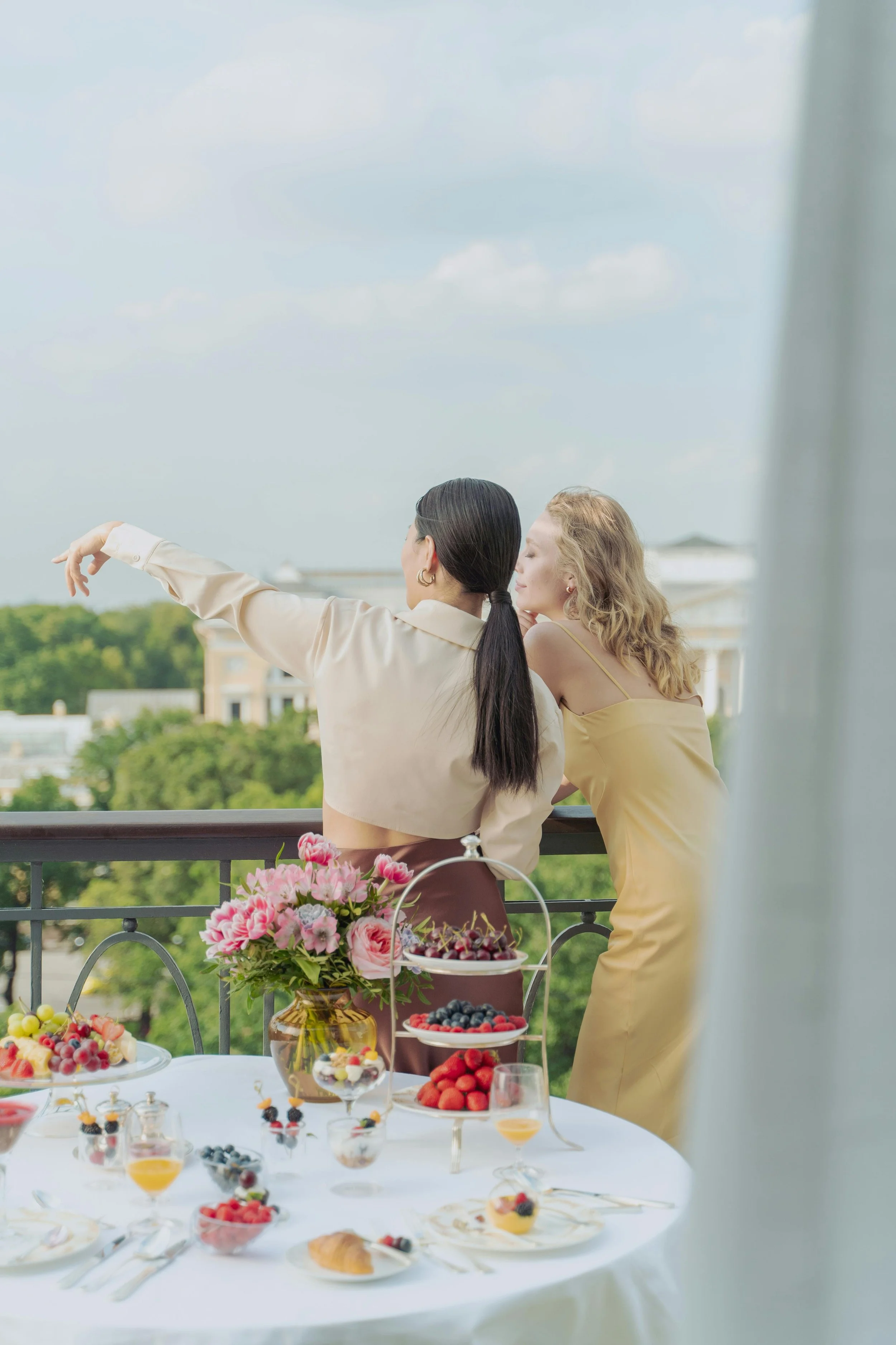 Two women standing on a balcony overlooking trees and buildings, one pointing at the distance, with a table of cakes, berries, and flowers in front of them.