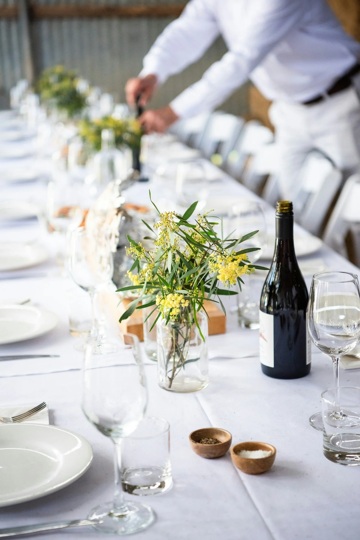 Table set for a meal with white plates, wine glasses, a wine bottle, a glass jar with yellow flowers, and small bowls of salt and pepper, with a person in a white shirt in the background