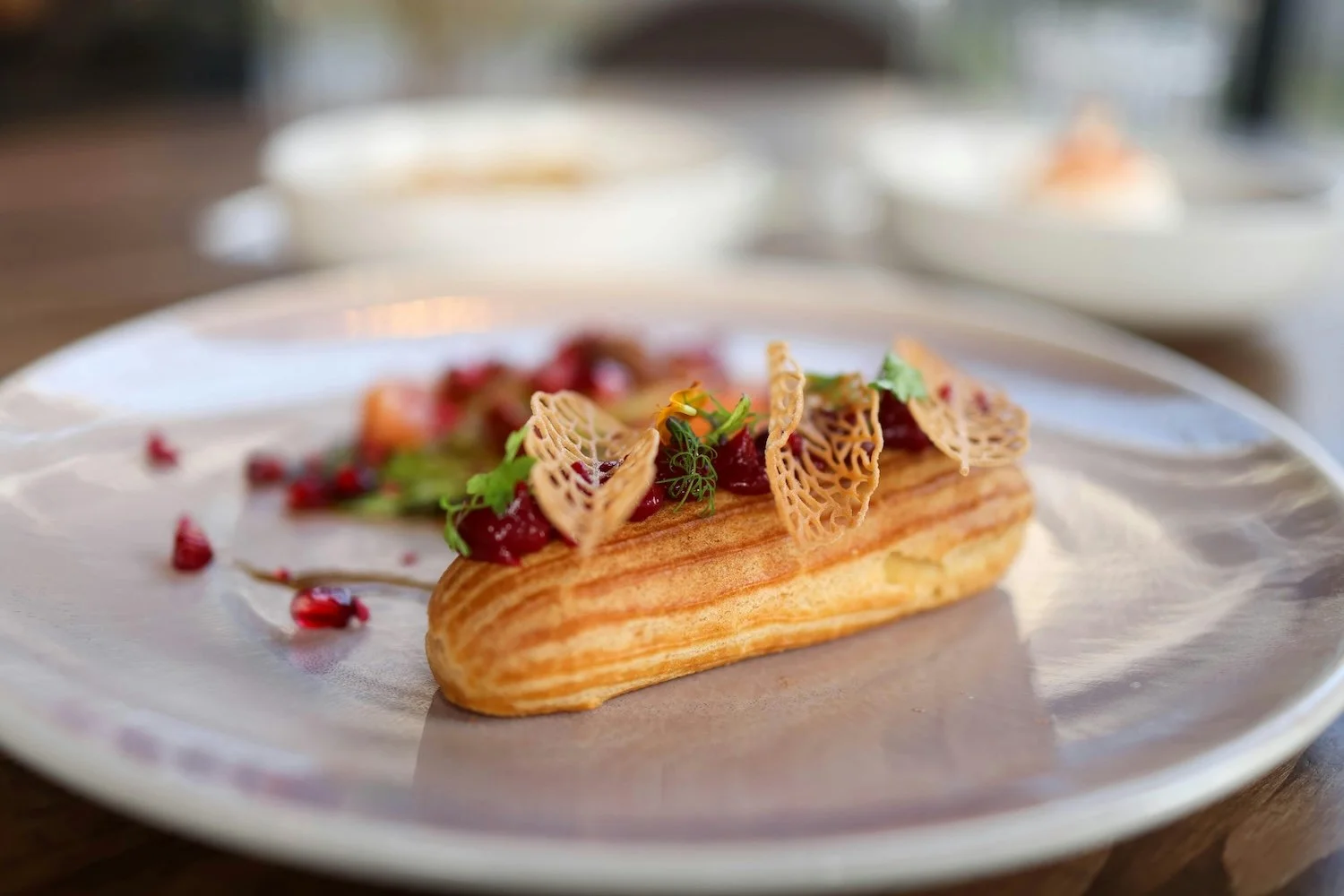Elegant plated éclair topped with berries, garnished with edible lacy structures and fresh herbs on a white ceramic plate.