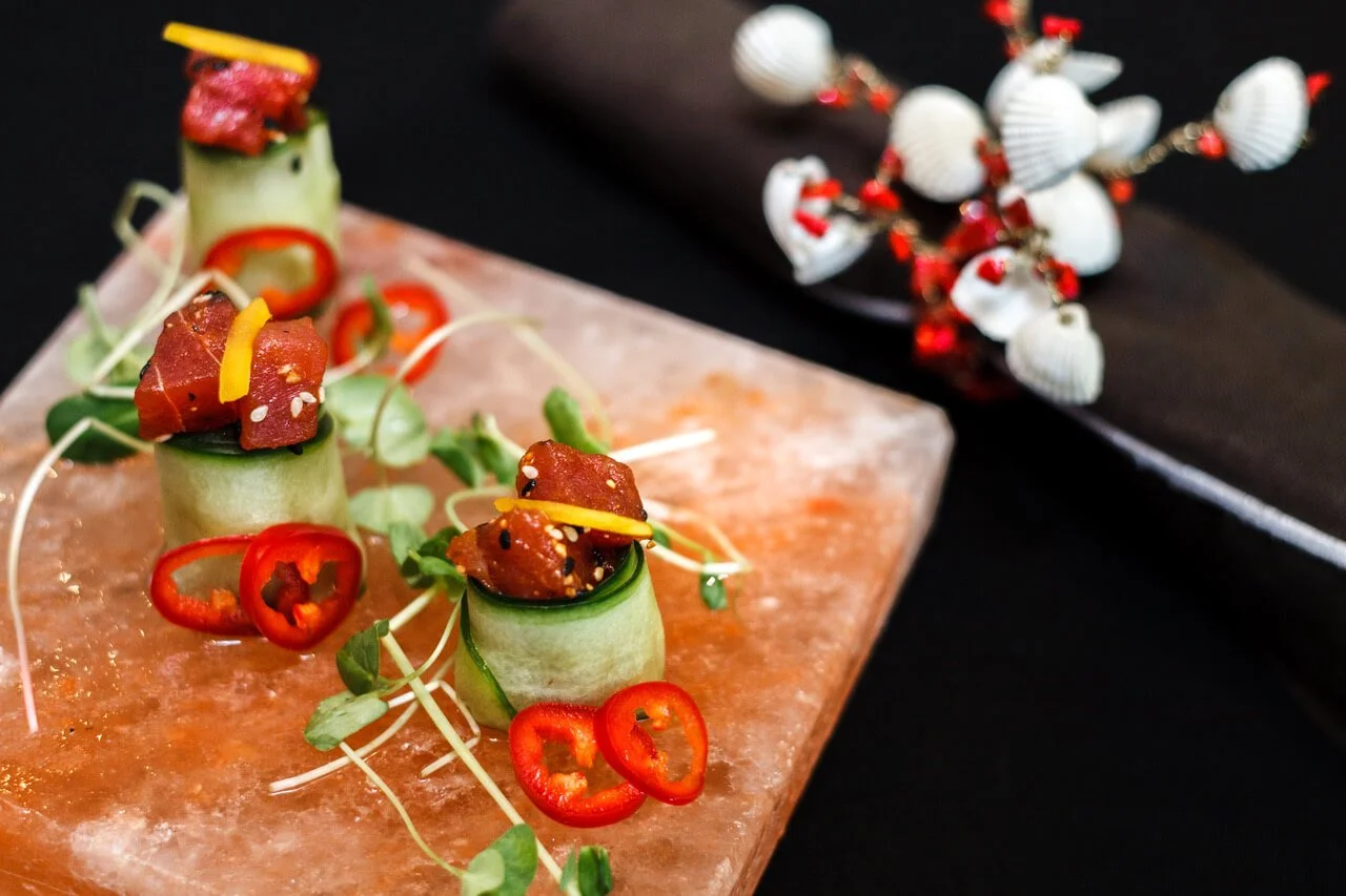 Cucumber rolls topped with raw tuna, garnished with red chili slices, sesame seeds, and microgreens, arranged on a salt block.