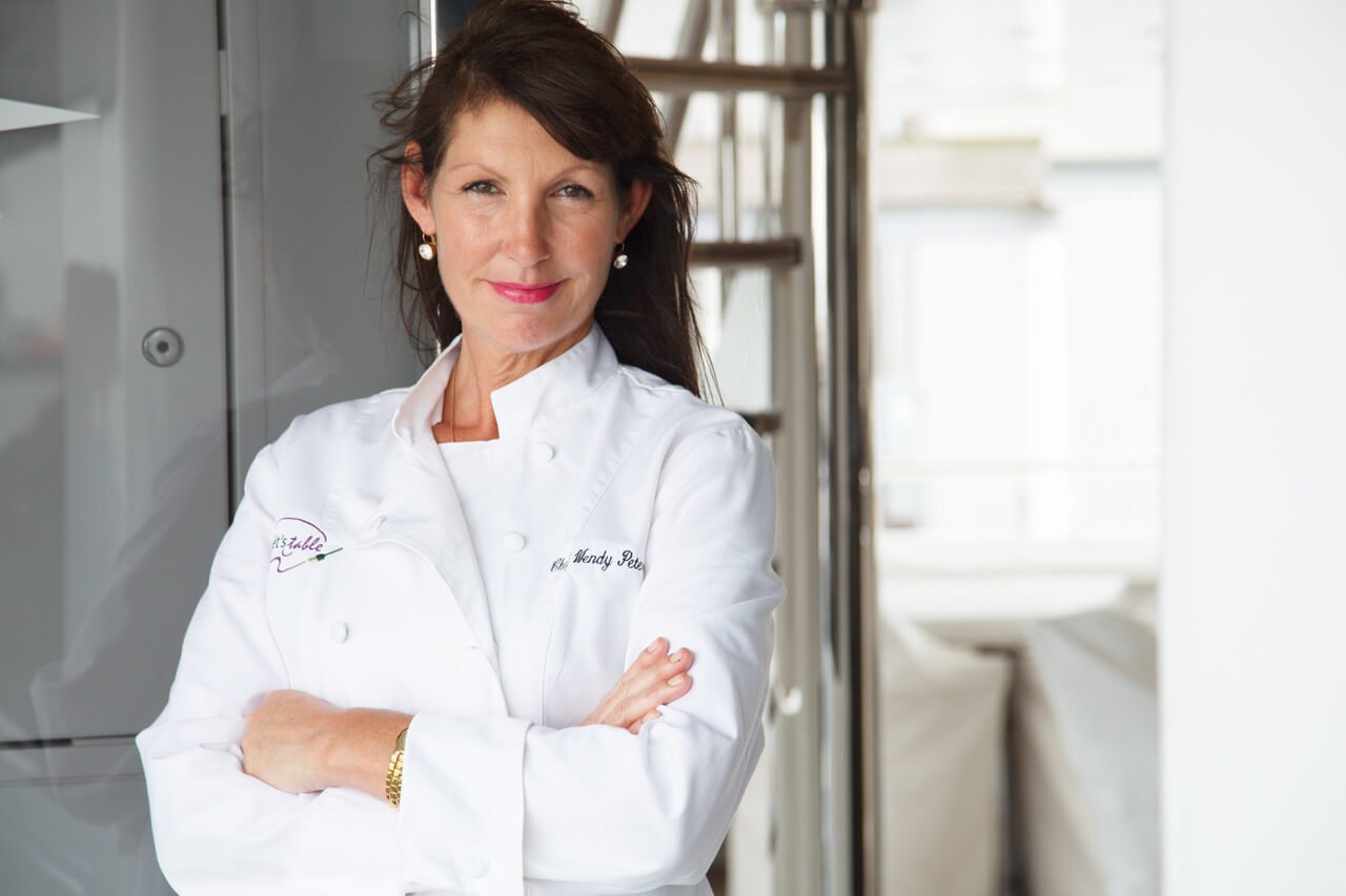 A woman chef with dark brown hair in a white chef's coat, standing with arms crossed in a professional kitchen, smiling at the camera.