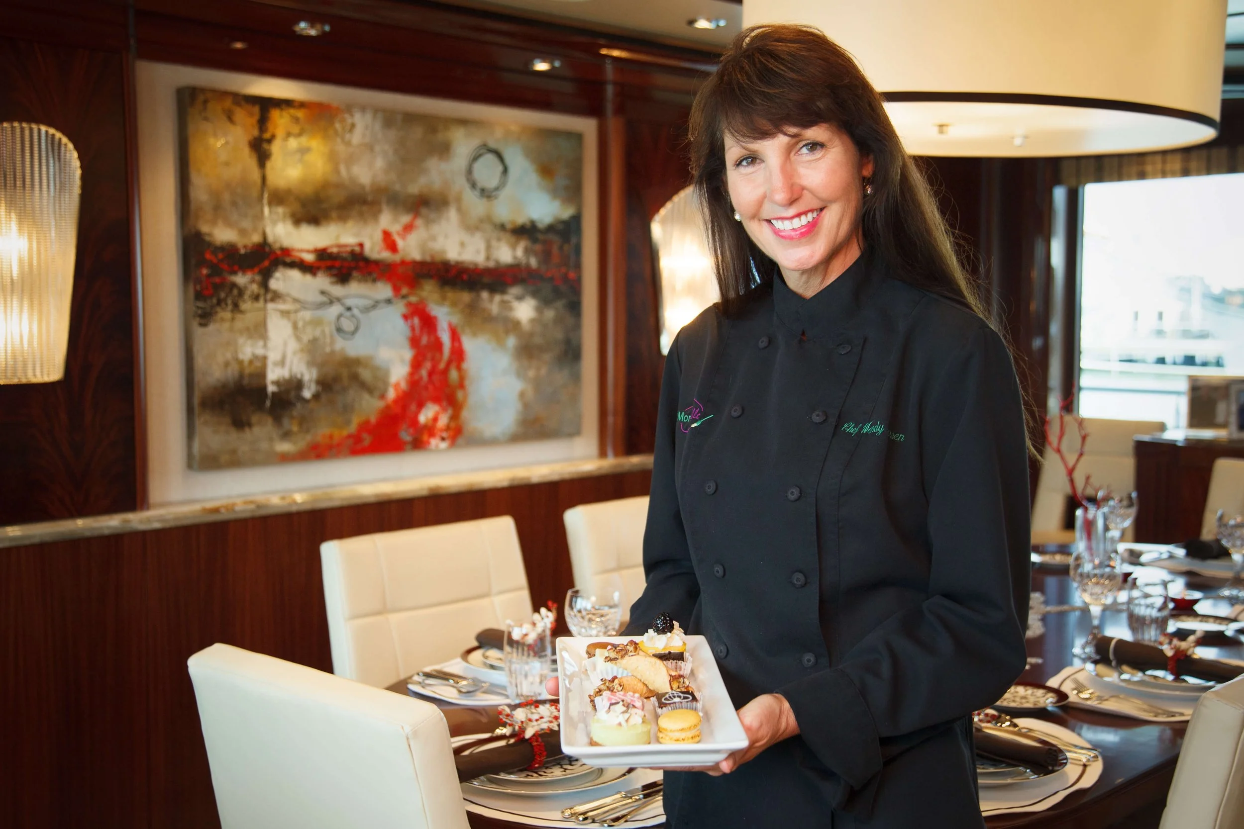 A woman in a black chef's jacket standing in a dining room holding a plate of assorted desserts, with a decorated dining table and abstract artwork in the background.