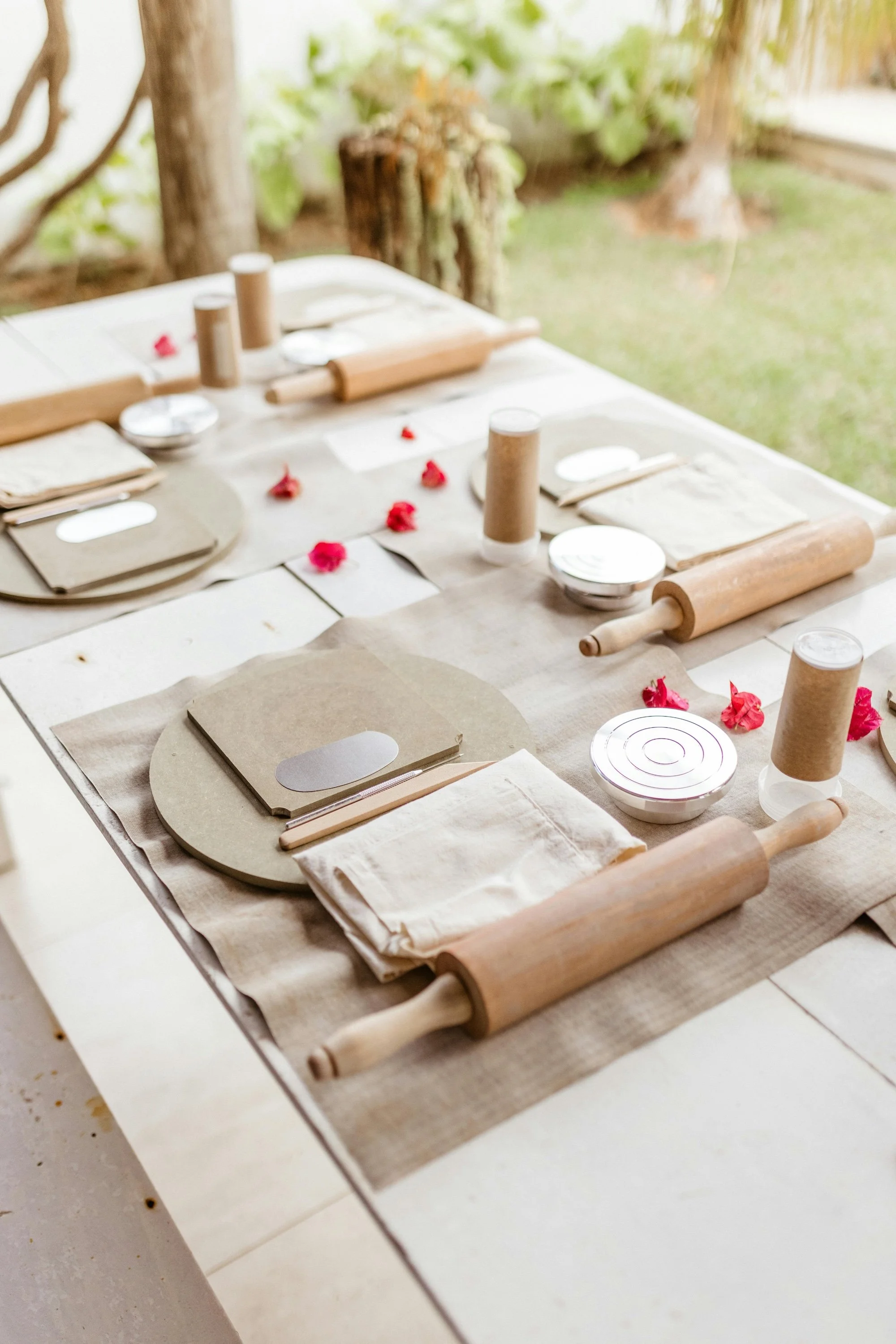 A tablescape for a baking or cooking activity, set with beige napkins, beige placemats, small bowls, rolling pins, and cutlery, decorated with scattered pink flower petals, outside with grass and trees in the background.