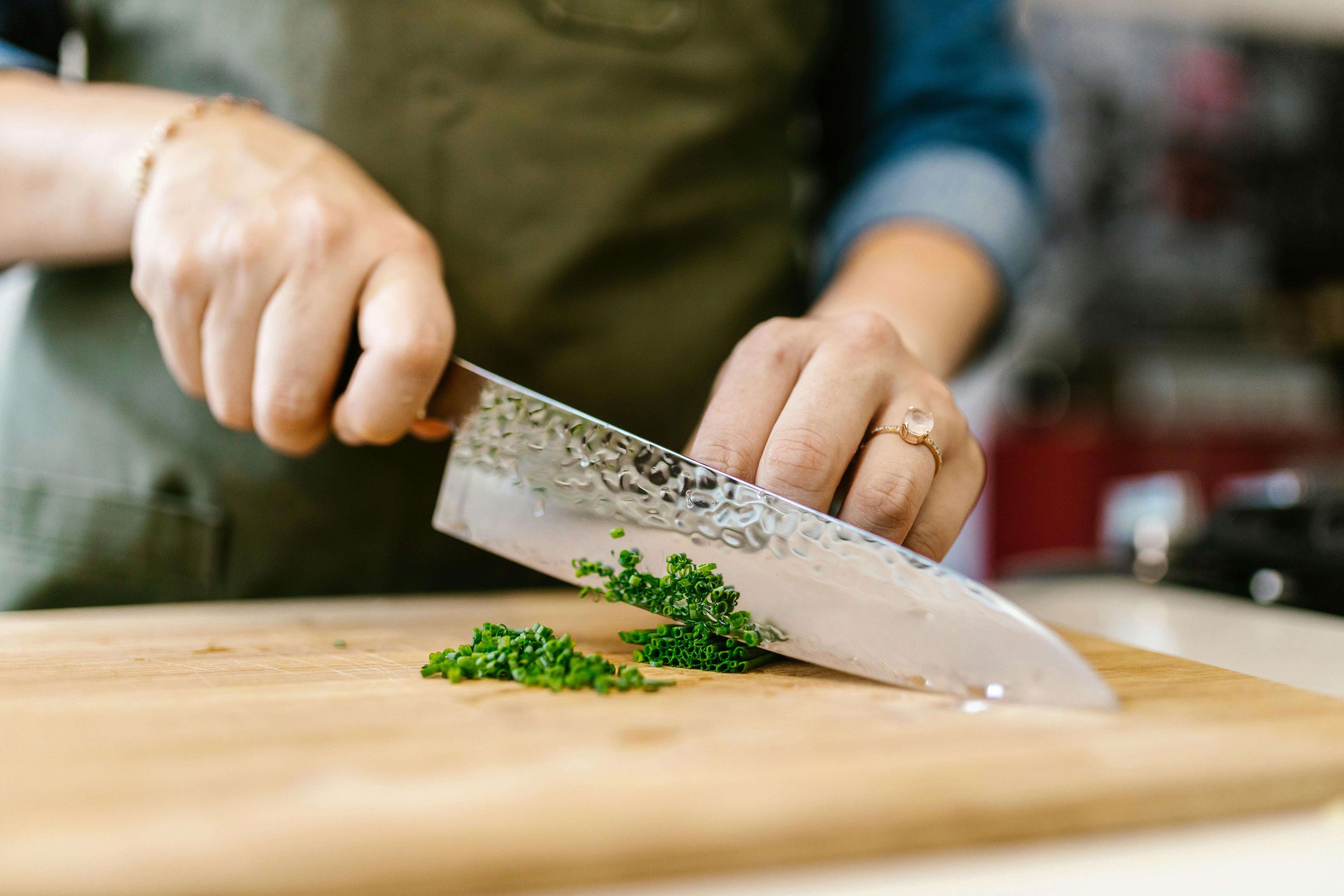 Person chopping green chives on a wooden cutting board using a textured metal knife.