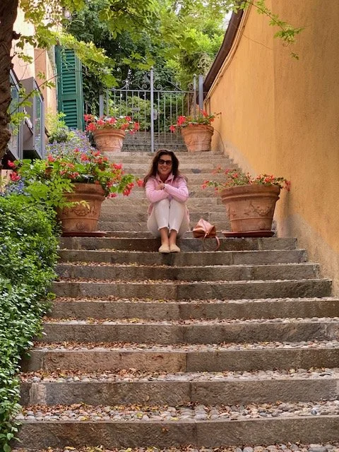 A woman sitting on stone steps outdoors, surrounded by potted flowers, with greenery and a gate in the background.