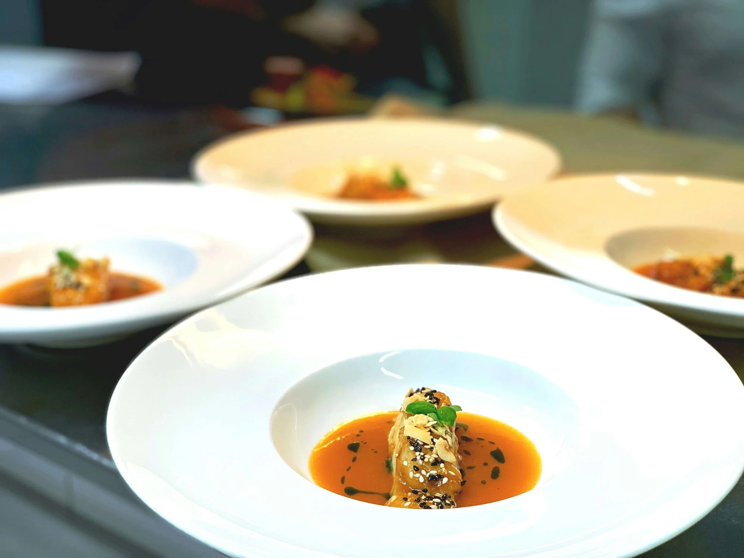 Close-up of a plated appetizer featuring a rolled dish garnished with greens and sesame seeds, served with a reddish sauce, on white plates.