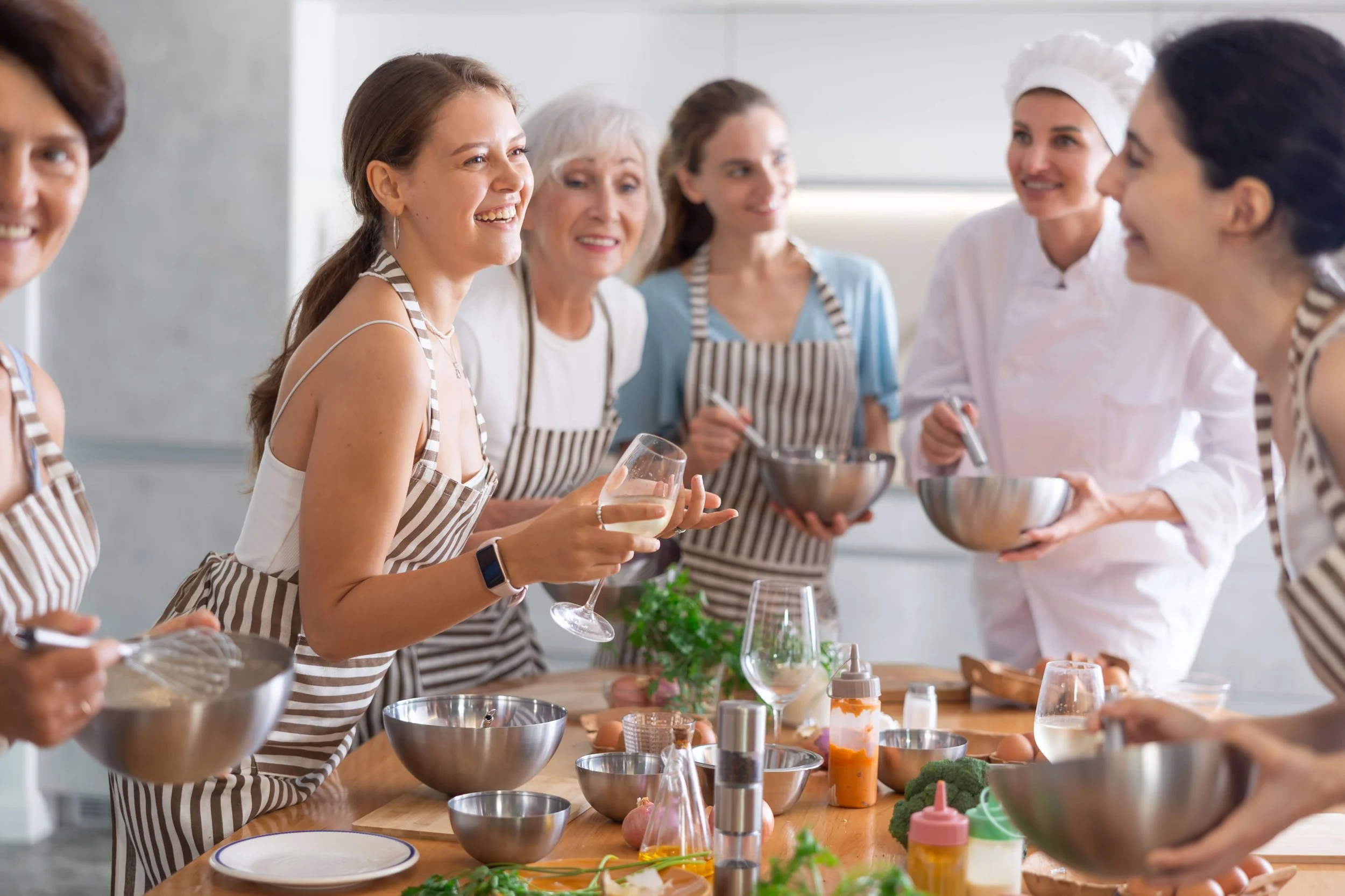 Women wearing aprons around a kitchen table, smiling and talking, with cooking ingredients and utensils.