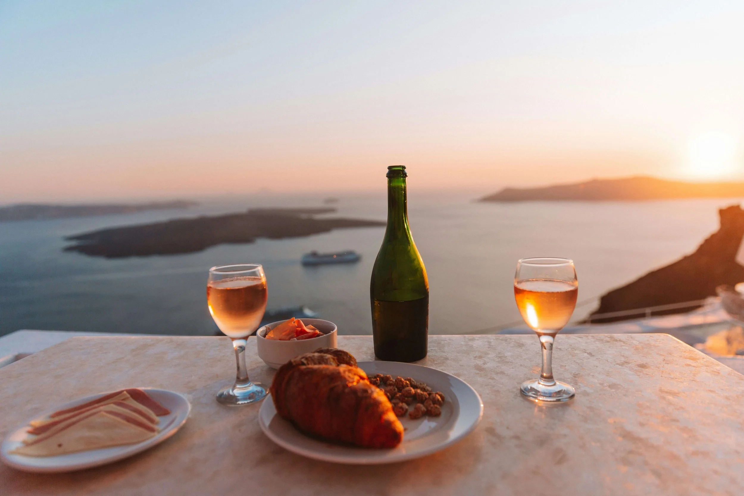 Dining table with a view of the ocean at sunset, featuring a bottle of wine, two glasses of rosé, a plate with meat, a bowl of salad, and a dish with sliced cheese.