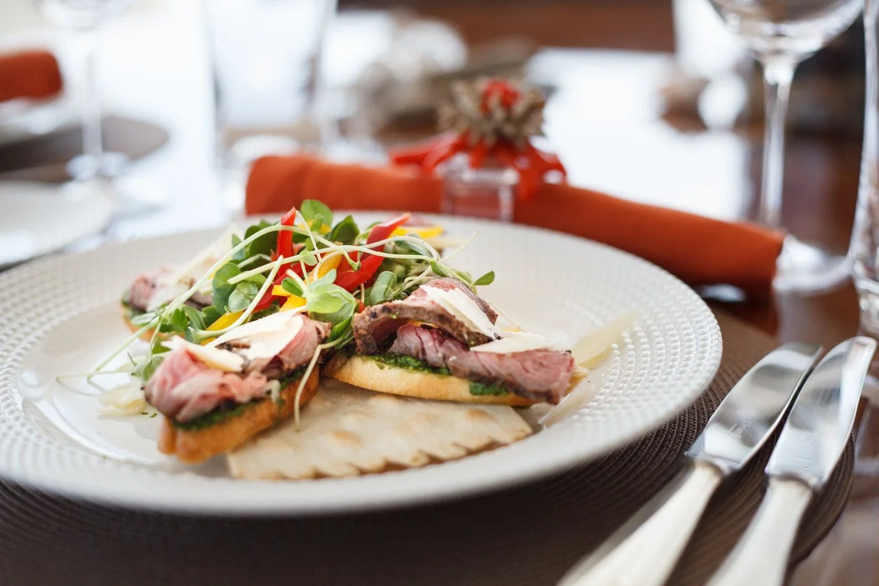 A plated meal featuring sliced beef on toasted bread with greens and microgreens, served on a white decorative plate. In the background, there are wine glasses and a rolled napkin with a small floral decoration.