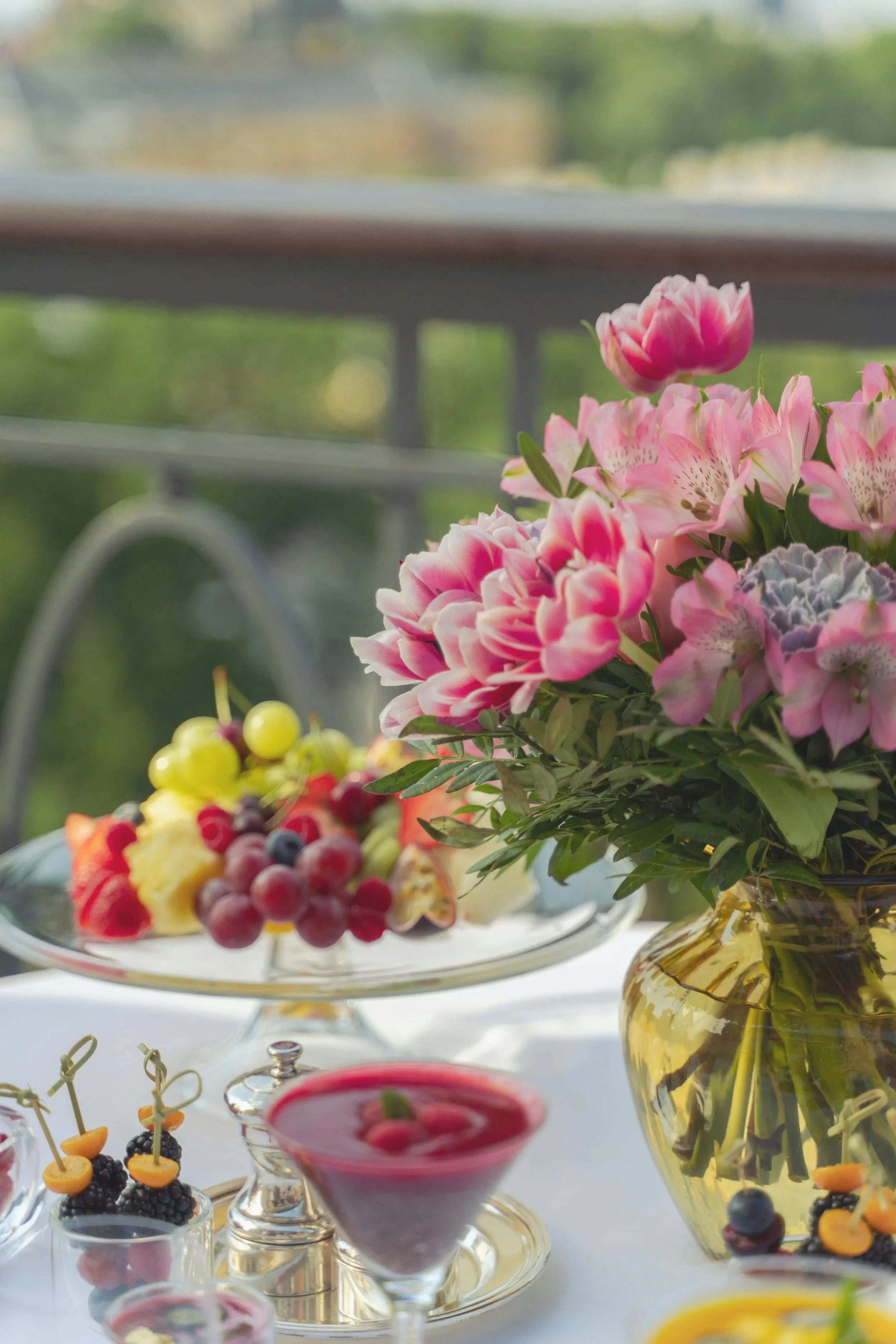 A floral centerpiece with pink and purple flowers in a yellow glass vase on a table, with assorted fruit and desserts, including a fruit platter with grapes and berries, and pink and purple berry desserts, set against a blurred outdoor background.