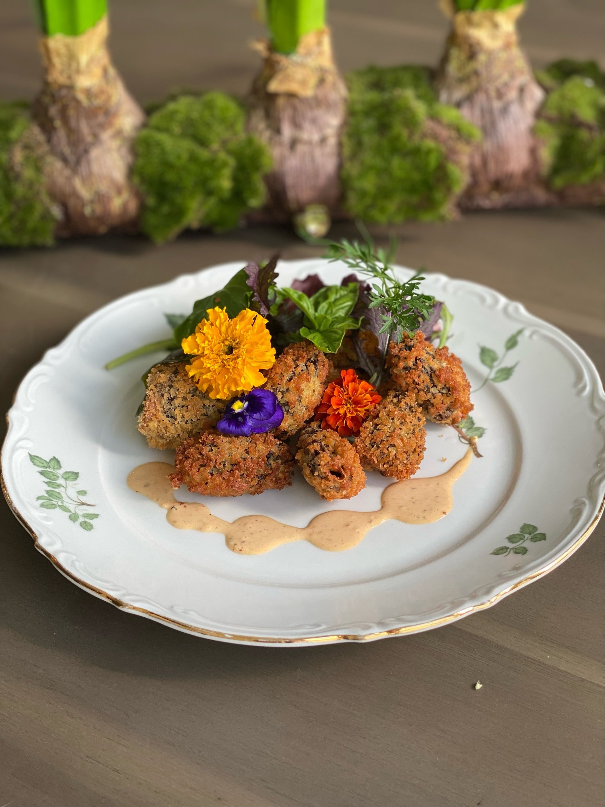 Fried chicken tenders garnished with edible flowers and herbs, served with a creamy dipping sauce on a white plate with green leaf patterns, with artichoke plants in the background.