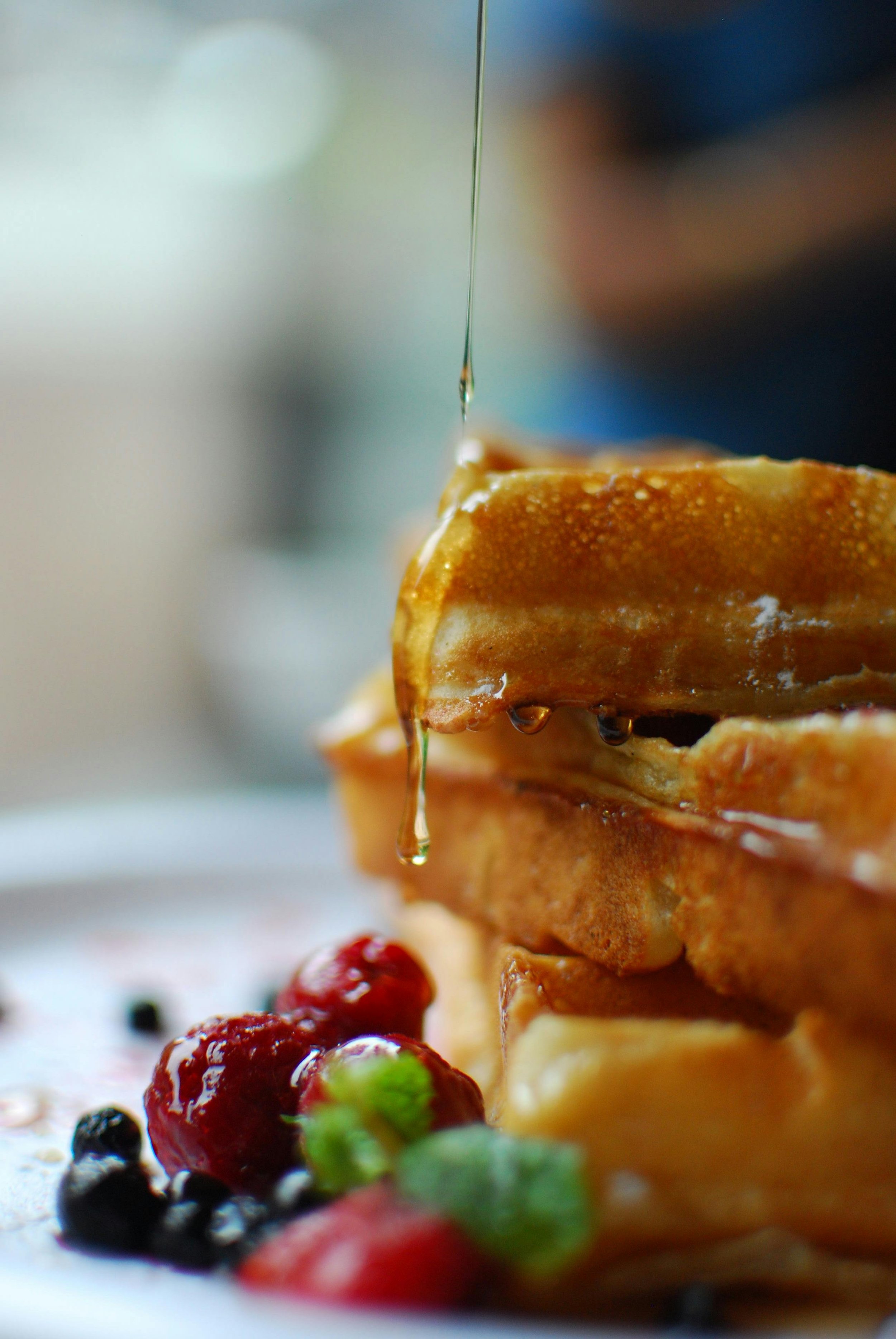 Close-up of golden brown waffles stacked with syrup dripping down, garnished with assorted berries including strawberries, blueberries, and raspberries.