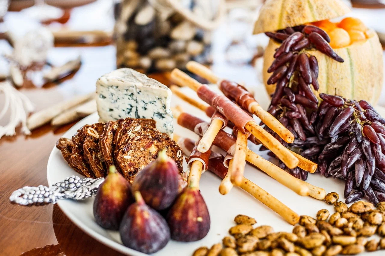 A white plate with cheeses, figs, crackers, breadsticks, and grapes on a wooden table.