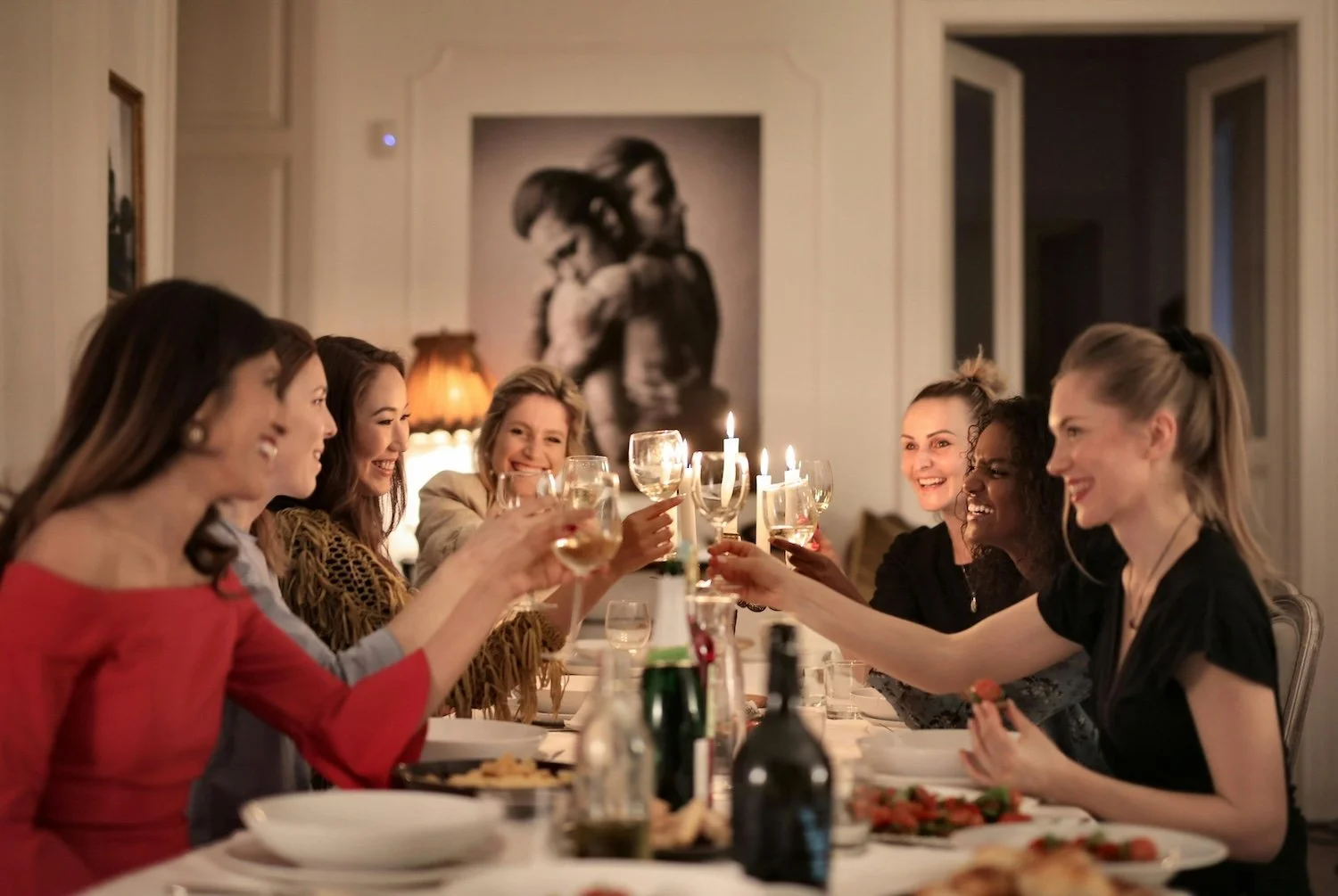 Group of women enjoying a dinner party, raising glasses in a toast, with candles and a painting in the background.