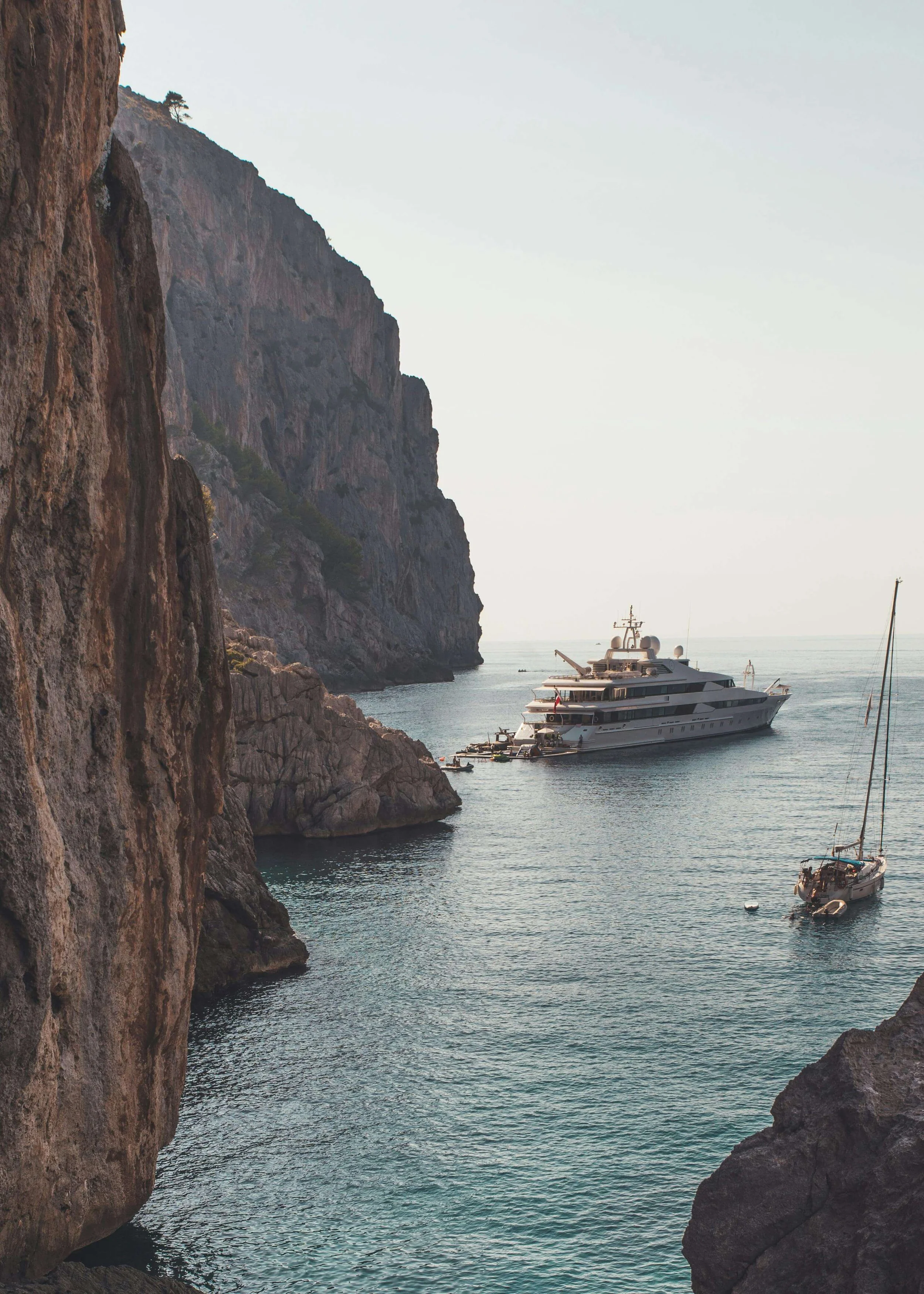 A large yacht and a sailboat anchored in a cove with rocky cliffs on one side and the open ocean on the other, under a clear sky.