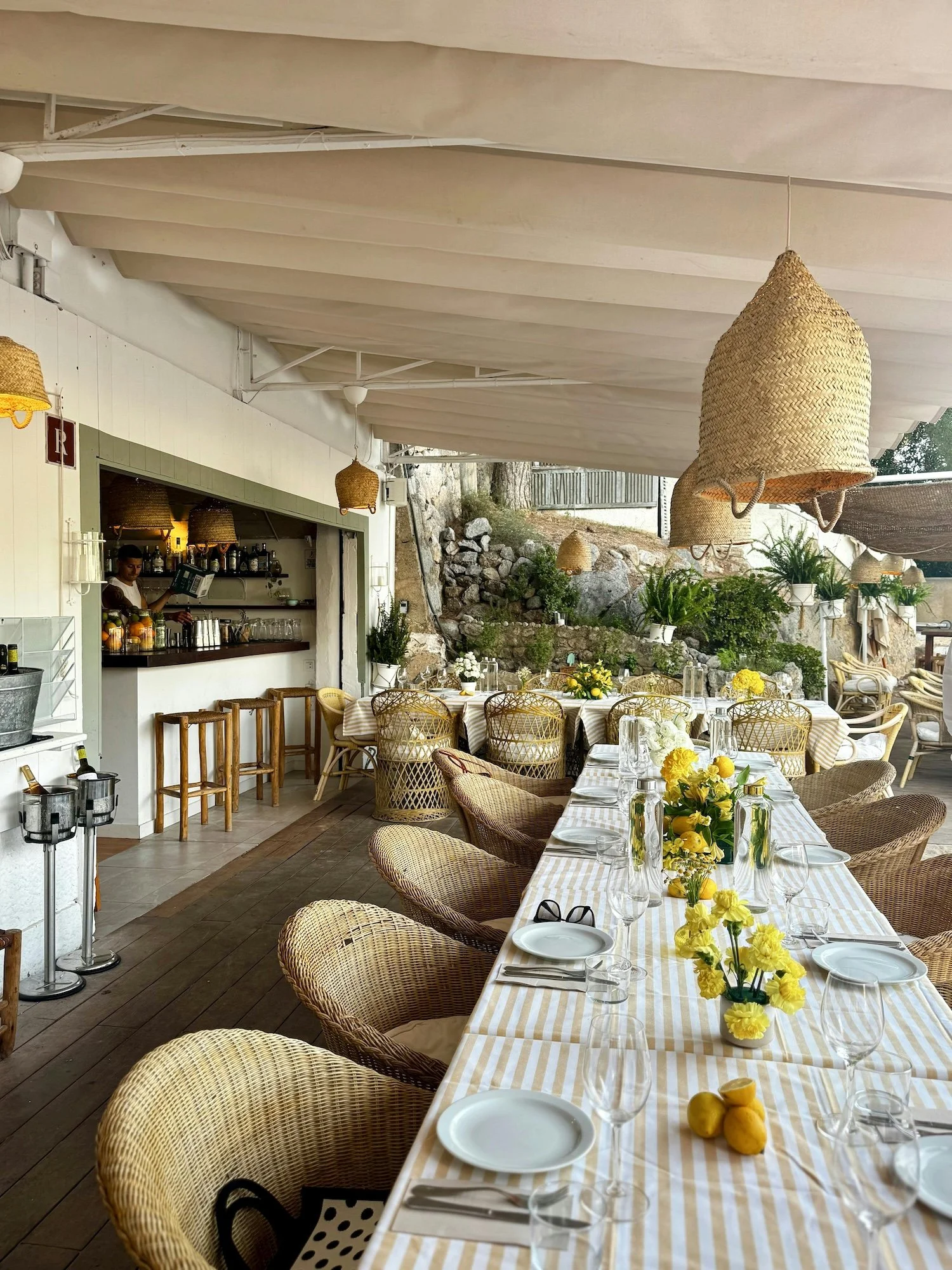 Outdoor dining area with a long table set with white plates, wine glasses, and yellow floral centerpieces, surrounded by wicker chairs and hanging wicker lights.