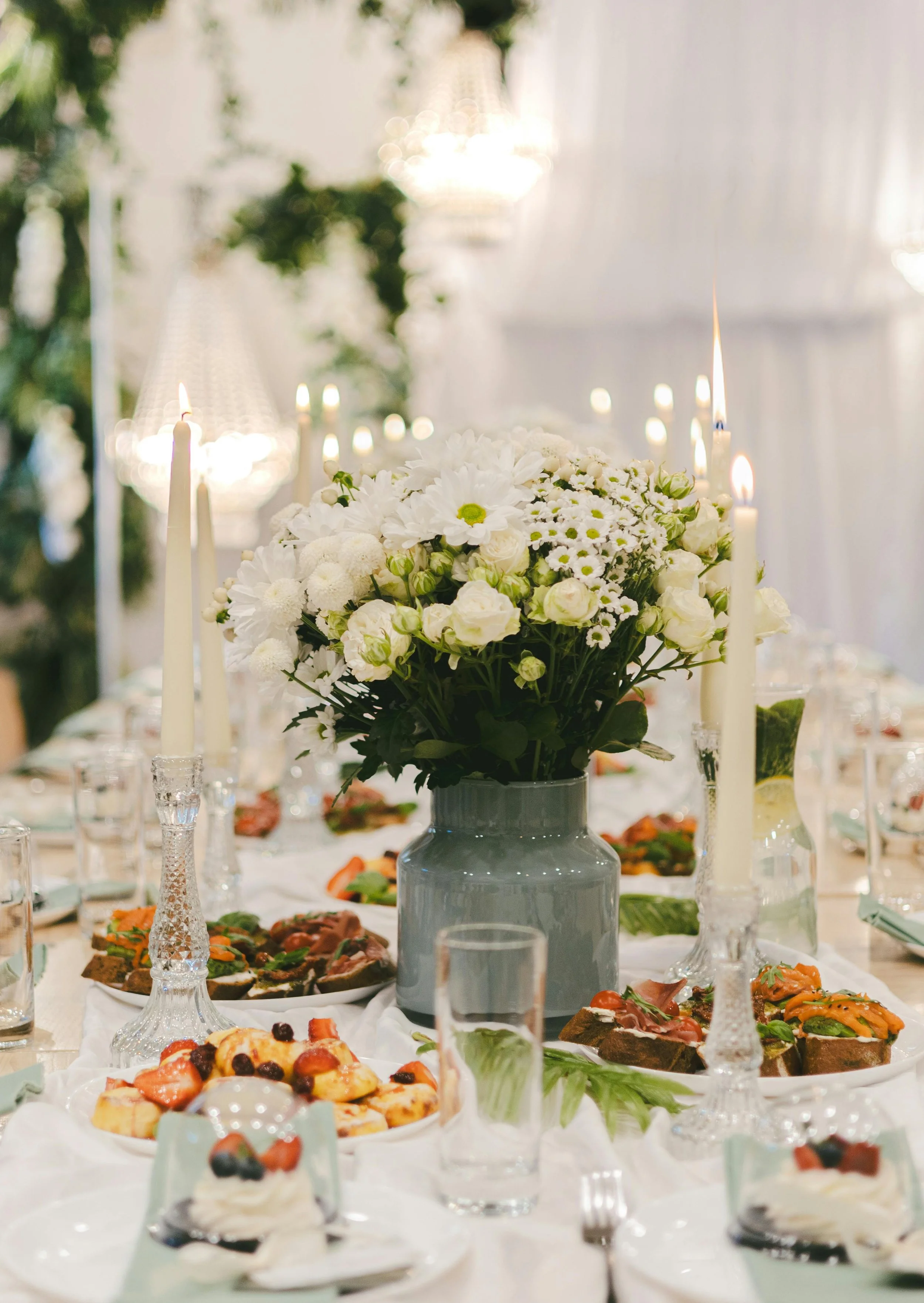 A banquet table decorated with a large bouquet of white flowers in a gray vase, surrounded by white candles in glass candlesticks, and topped with various dishes including desserts and appetizers, set in an elegant dining setting with chandeliers and