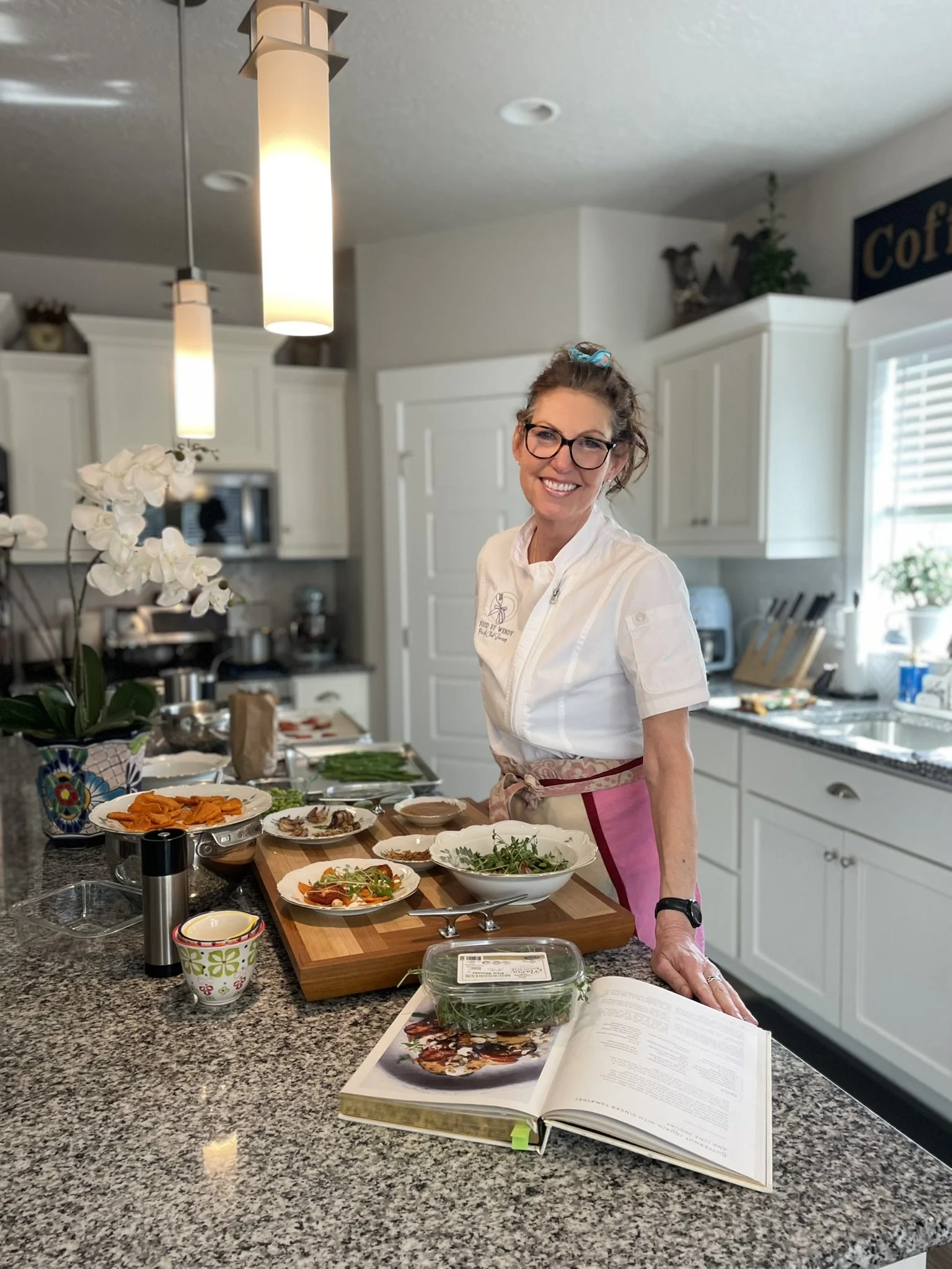 A woman in a white chef's jacket and apron, smiling, standing in a modern kitchen surrounded by prepared ingredients and dishes on a countertop.