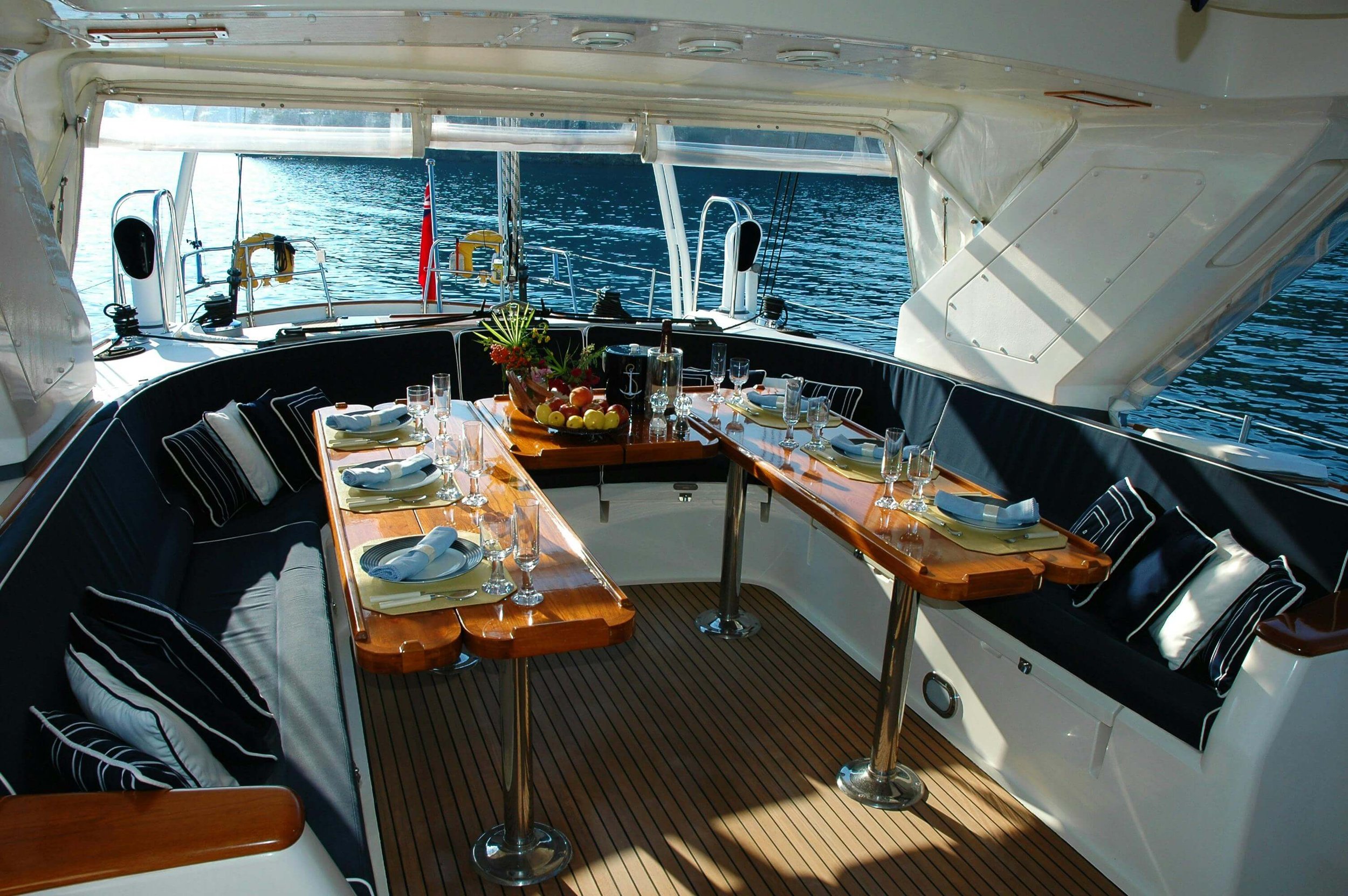 A yacht deck set for dining with a U-shaped seating area, a wooden table, and place settings, overlooking calm blue water.