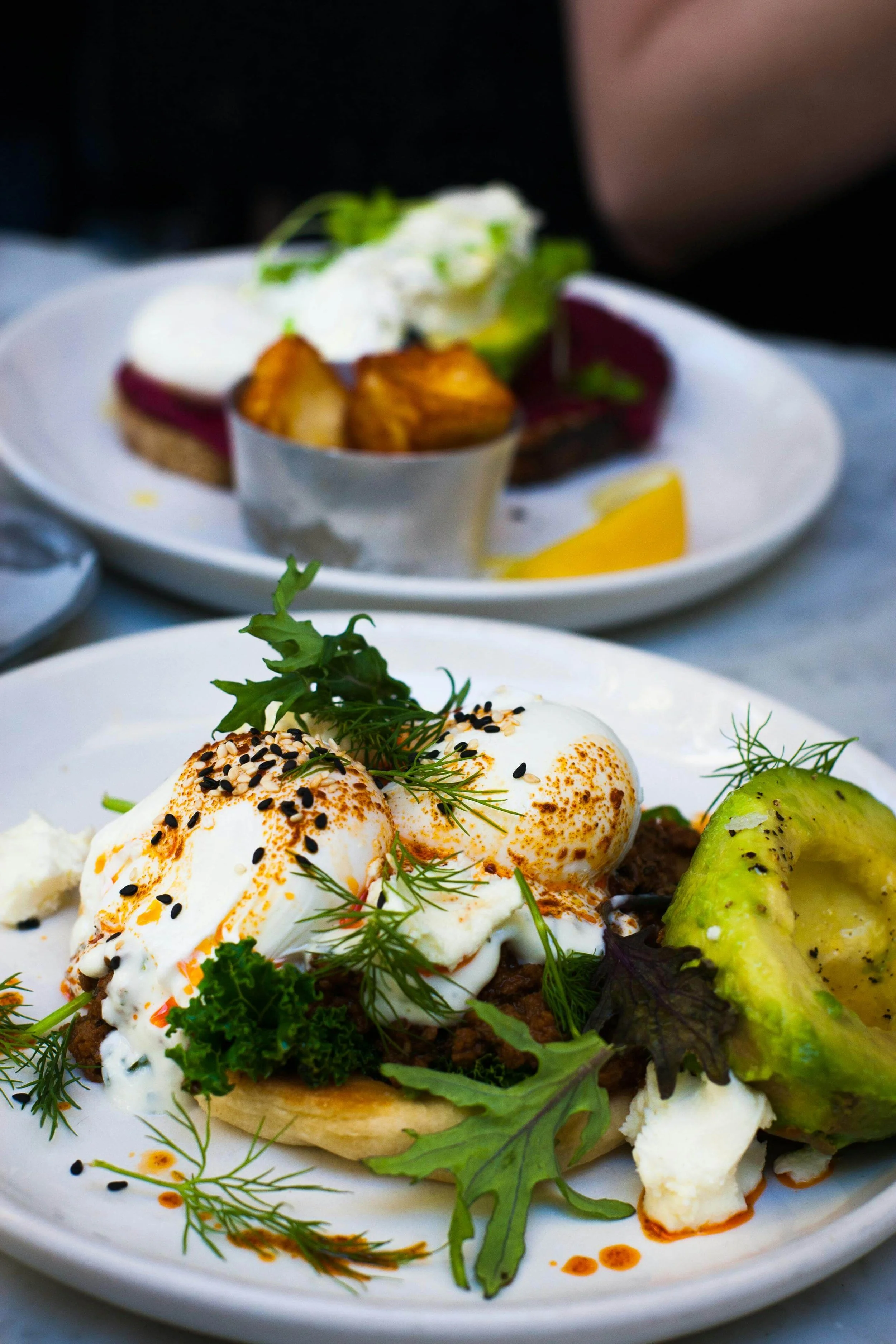 Close-up of a plate with poached eggs, avocado slices, greens, and seasonings, with another plate of food in the background.