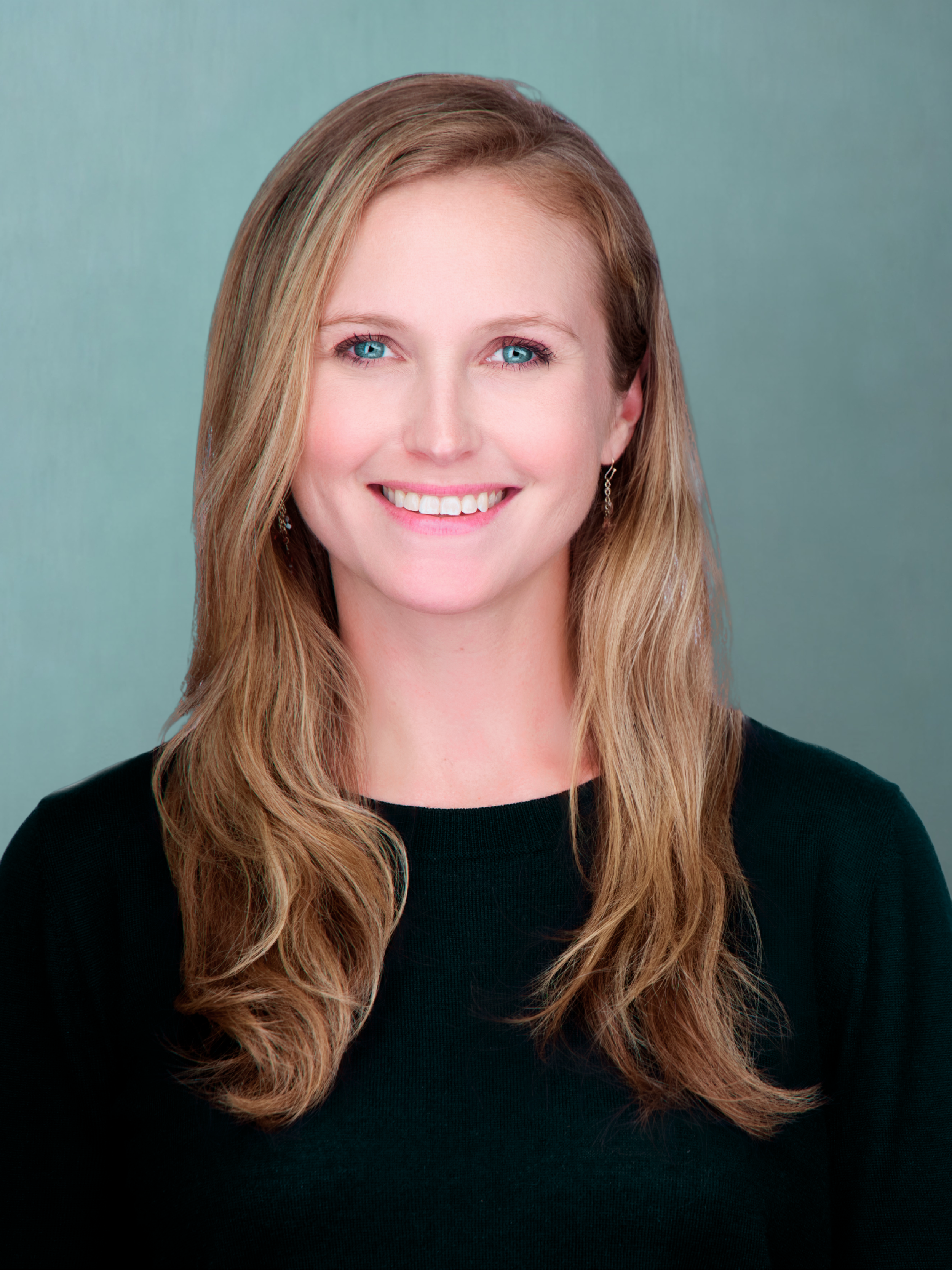 Close-up portrait of a woman with long, wavy blonde hair, wearing a black top, smiling against a soft teal background.