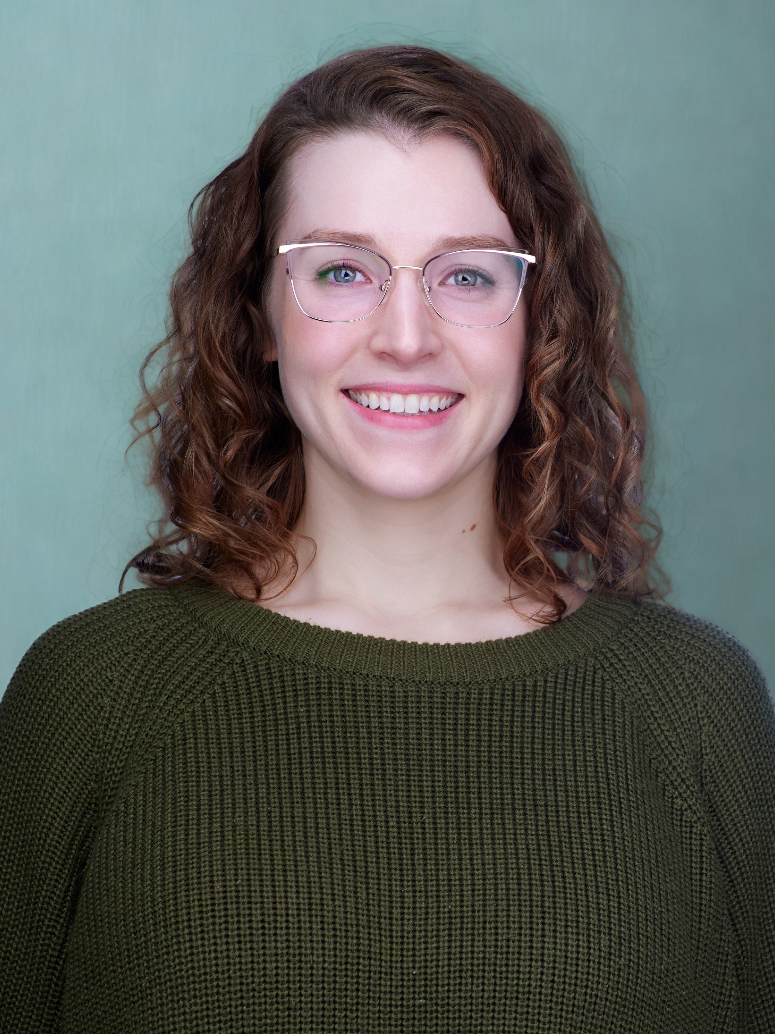 A woman with curly brown hair and glasses smiling at the camera, wearing a green sweater, standing against a light blue-green background.