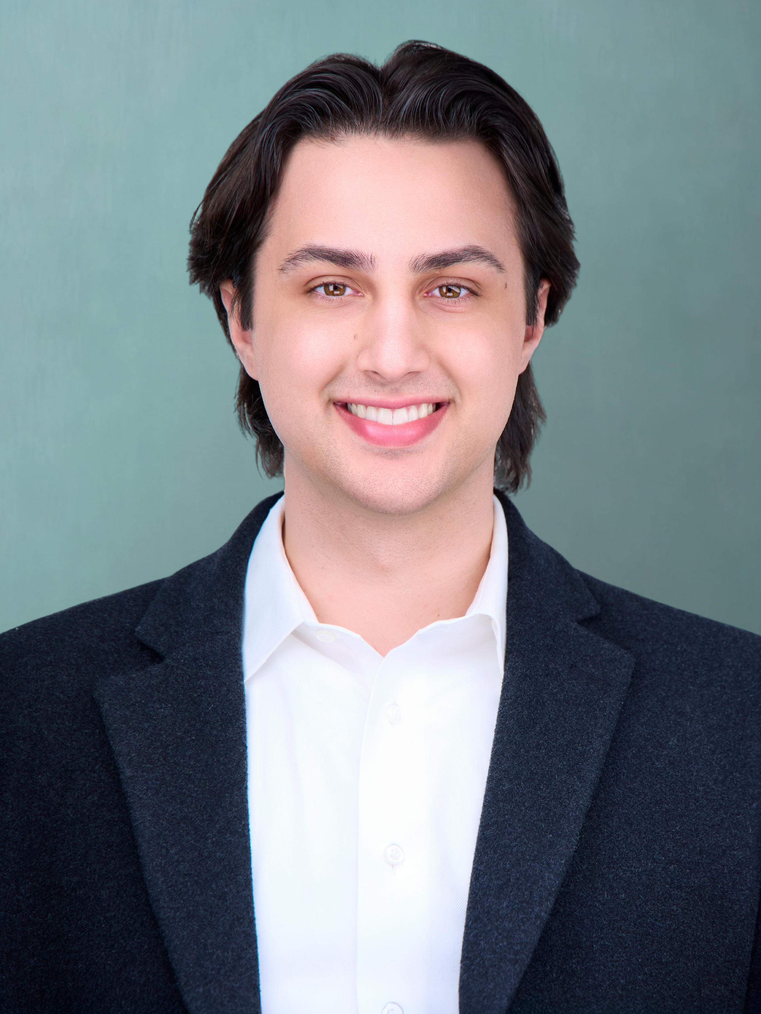 A young man with dark brown hair, brown eyes, wearing a white shirt and dark blazer, smiling against a plain teal background.