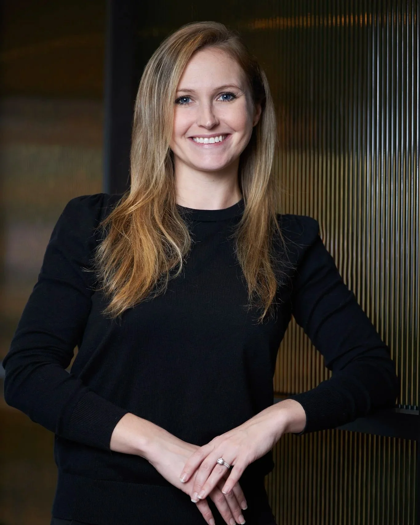 A young woman with long blonde hair, wearing a black top, smiling, standing indoors, leaning on a gold-colored striped wall panel.