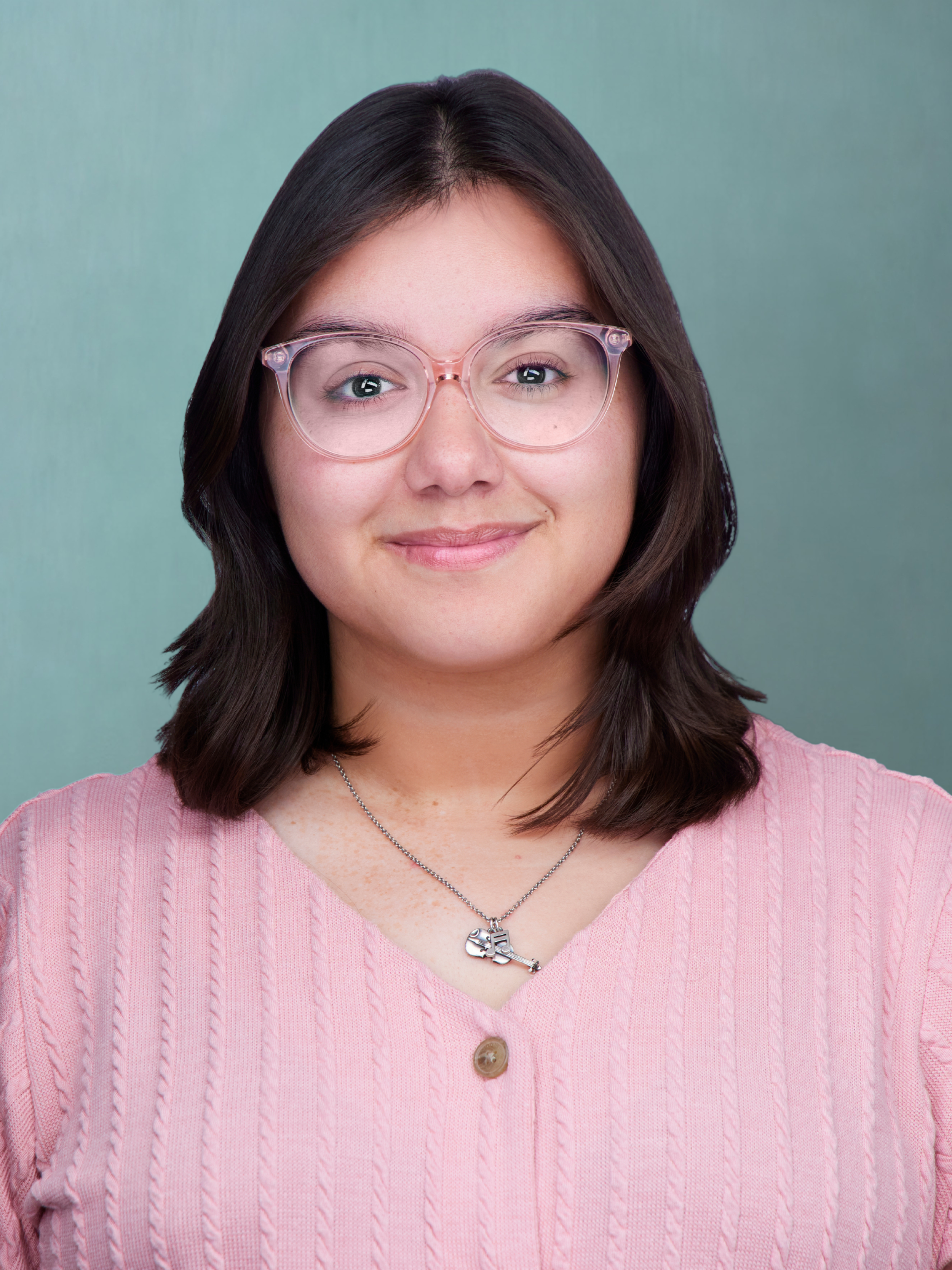 A woman with shoulder-length dark hair, wearing pink-framed glasses, a pink sweater with a cable knit pattern, and a necklace with a key-shaped pendant, standing against a teal background.