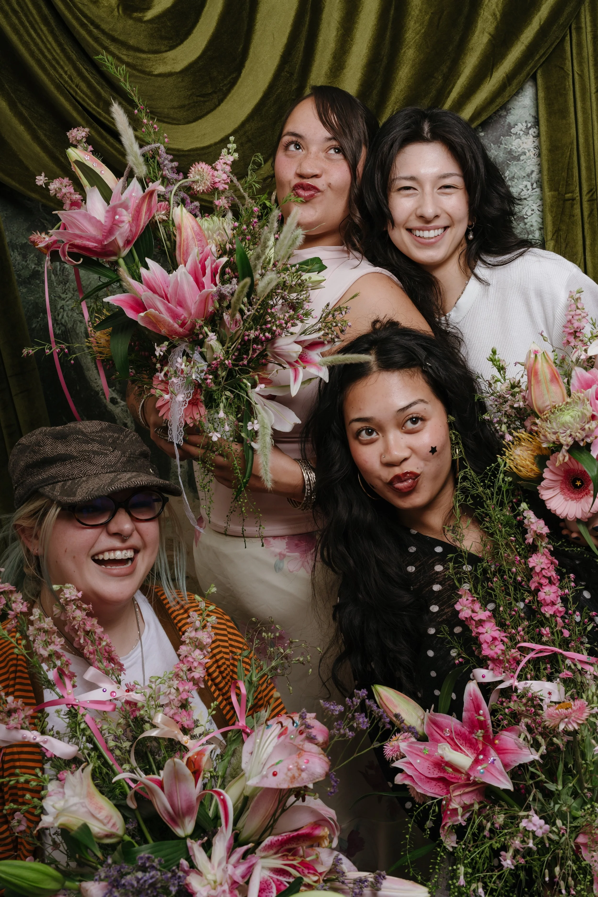 Group of diverse women smiling and posing with large bouquets of pink and white flowers indoors, with green velvet curtains in the background.