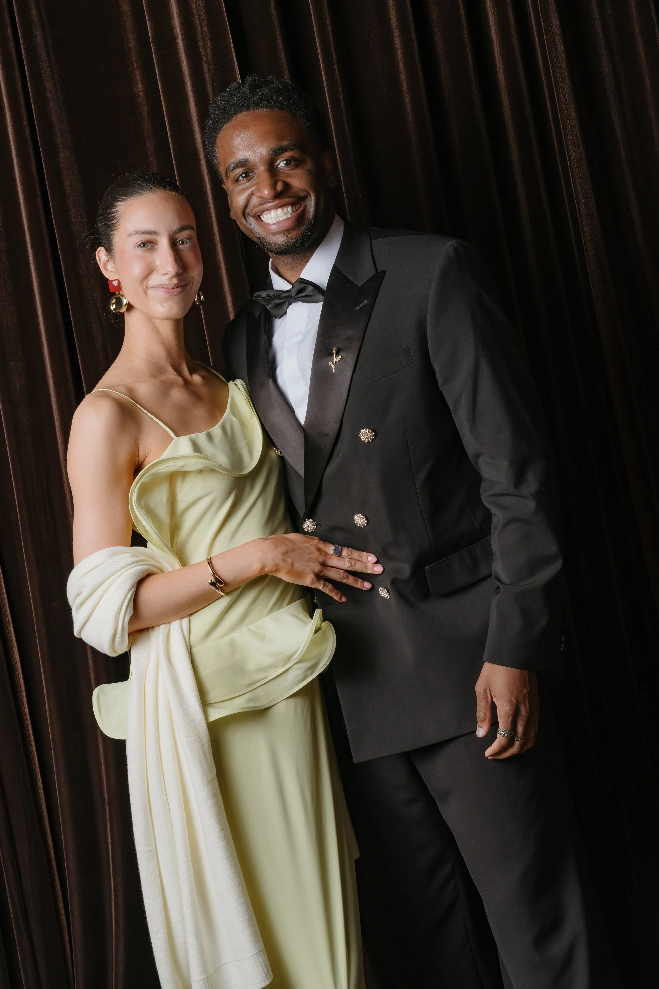 A smiling couple dressed in formal attire, standing close together in front of dark curtains.
