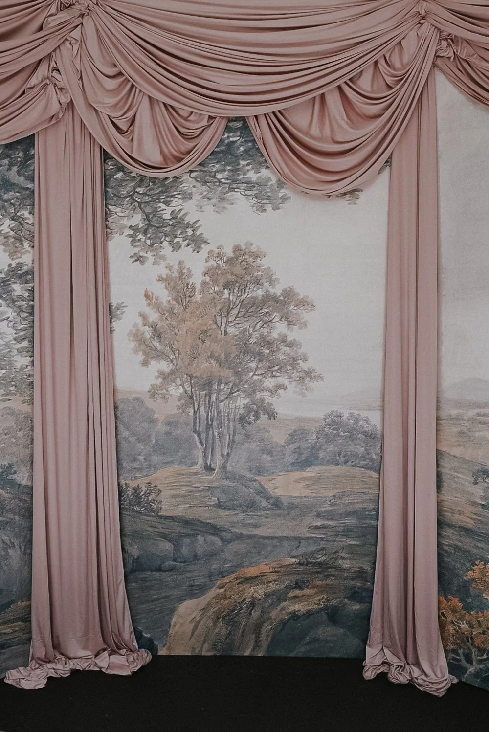 Fabric drapes in pink framing a wall mural of a landscape scene with trees and rolling hills.