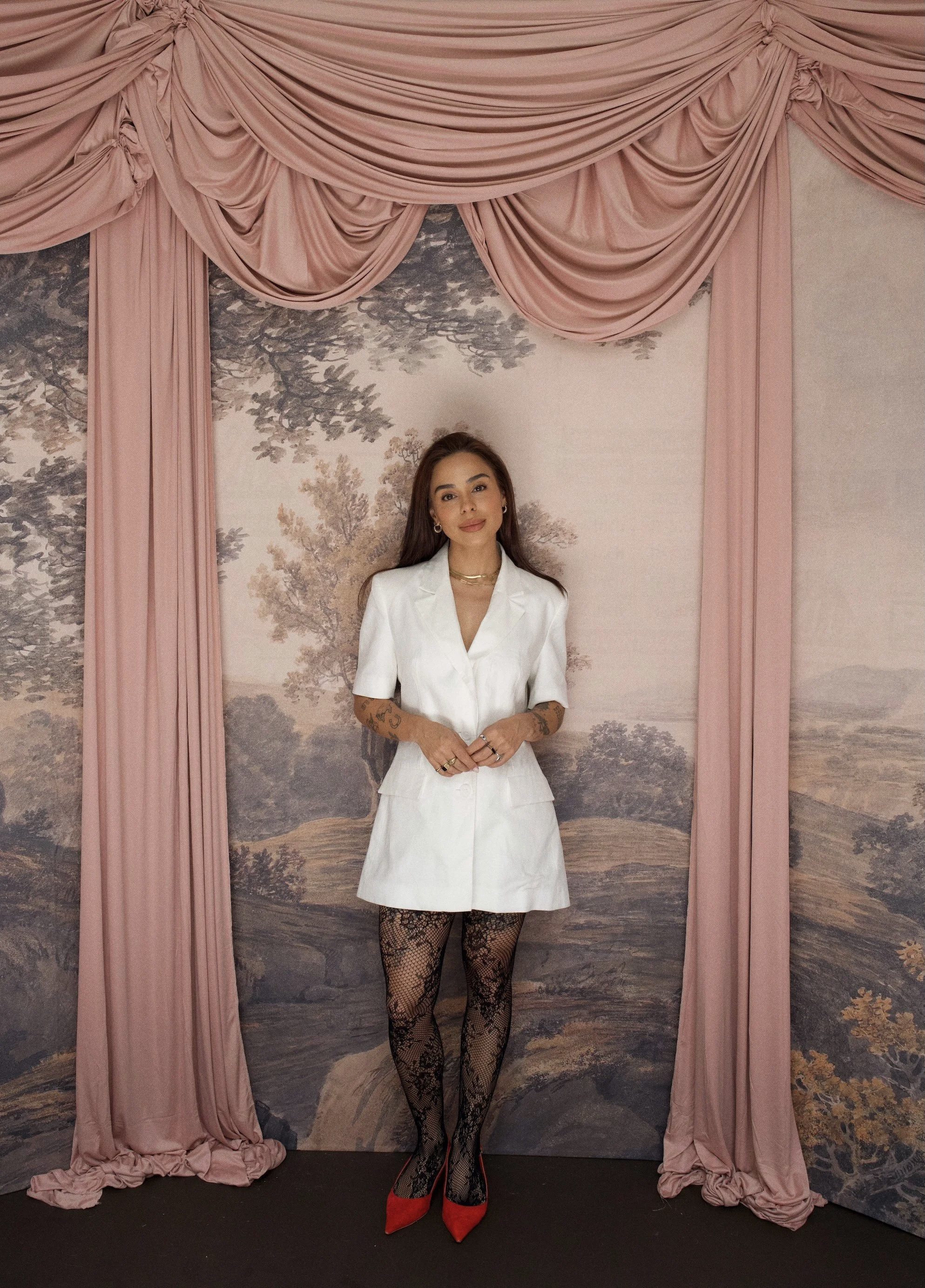 A woman with long brown hair and tattoos on her arms, dressed in a white outfit with black lace stockings and red heels, standing in front of a scenic wall with pink curtains and a landscape painting.