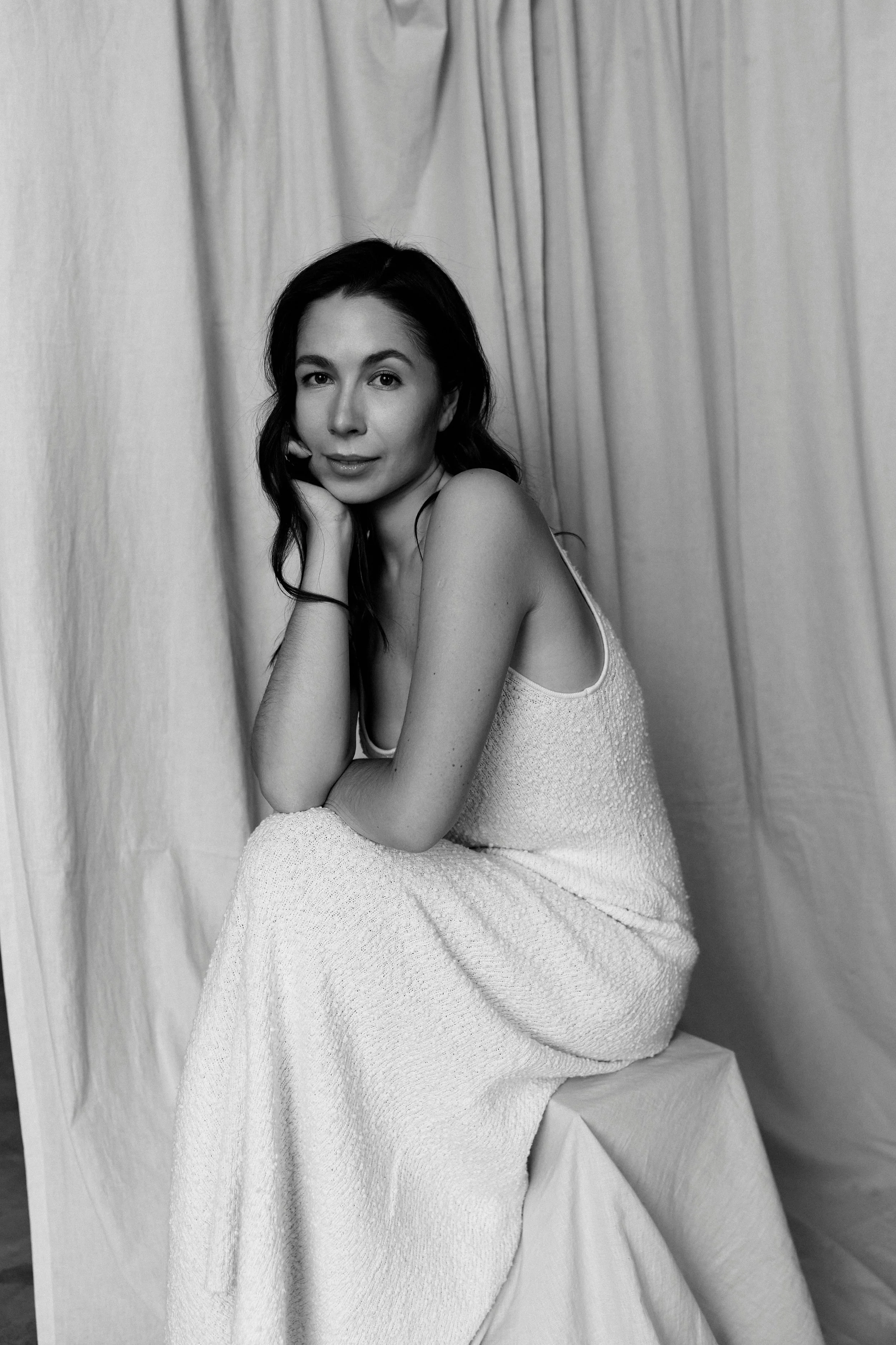 A black and white photograph of a woman with long dark hair, sitting on a stool, resting her chin on her hand, wearing a sleeveless, textured dress, with a curtain backdrop.