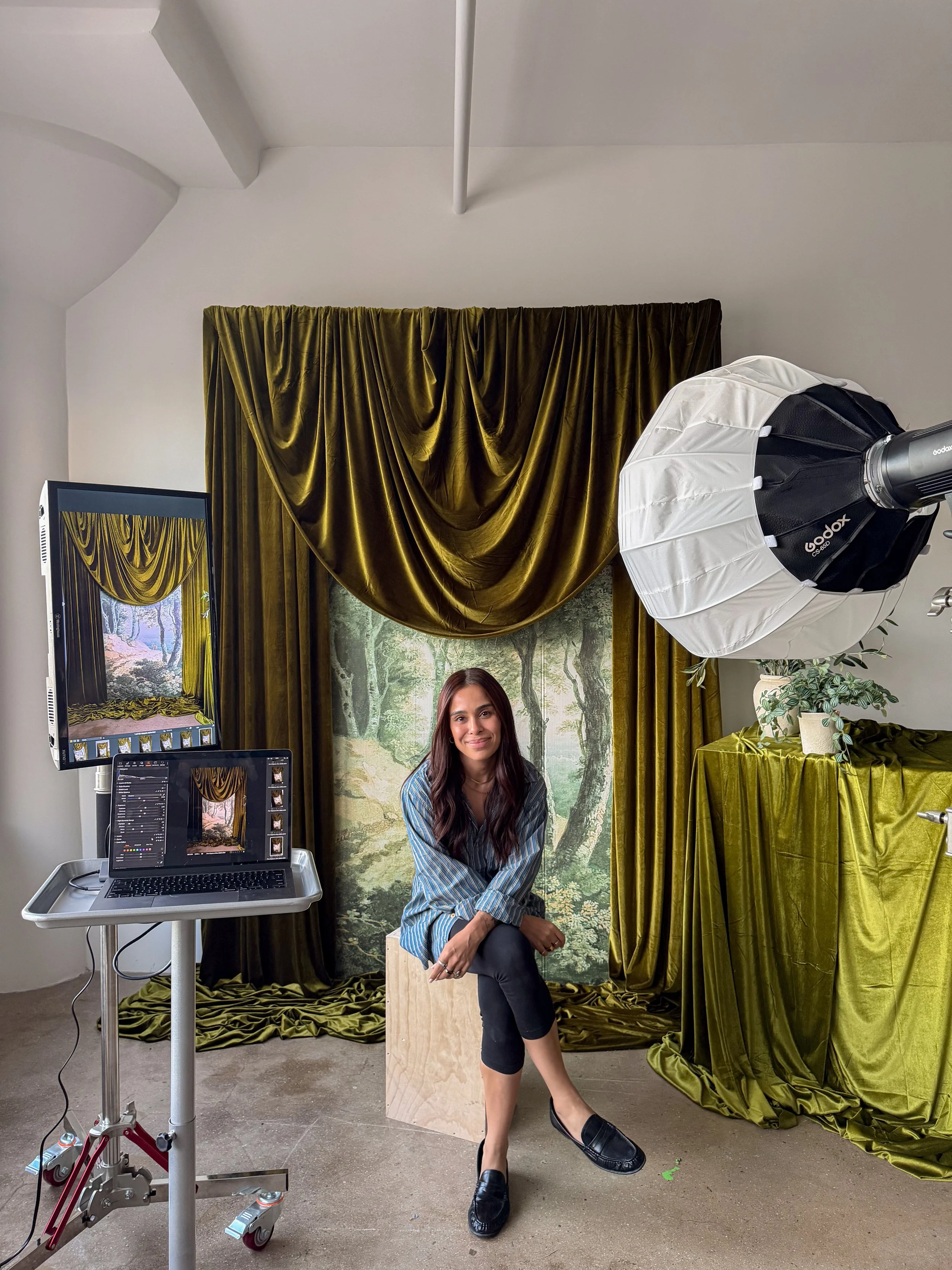 A woman with long dark hair sitting on a wooden box in a photography studio, posing with one foot crossed over the other and smiling. Behind her is a backdrop of a forest scene with green trees and a pathway, framed by large gold velvet curtains. To her left are photography equipment including a monitor, laptop, and a large softbox light.