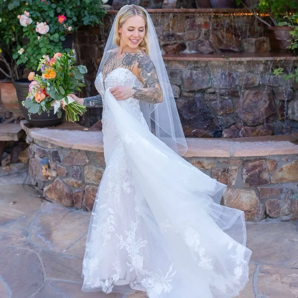 Bride in a white wedding gown with lace details, holding a colorful bouquet, standing outdoors near a stone waterfall feature, smiling and looking back over her shoulder.