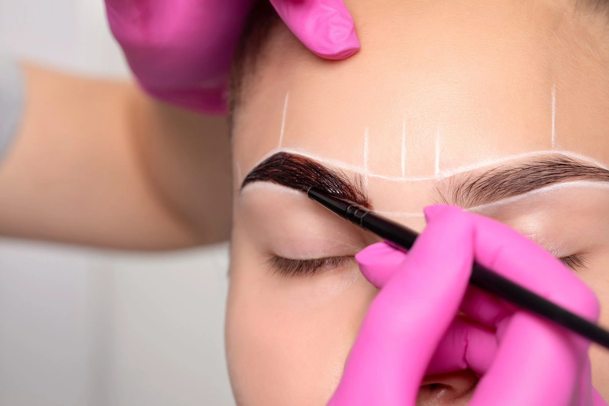A person with pink gloves applying makeup or cosmetic product to their eyebrow with a brush, with white chalk guidelines on the forehead