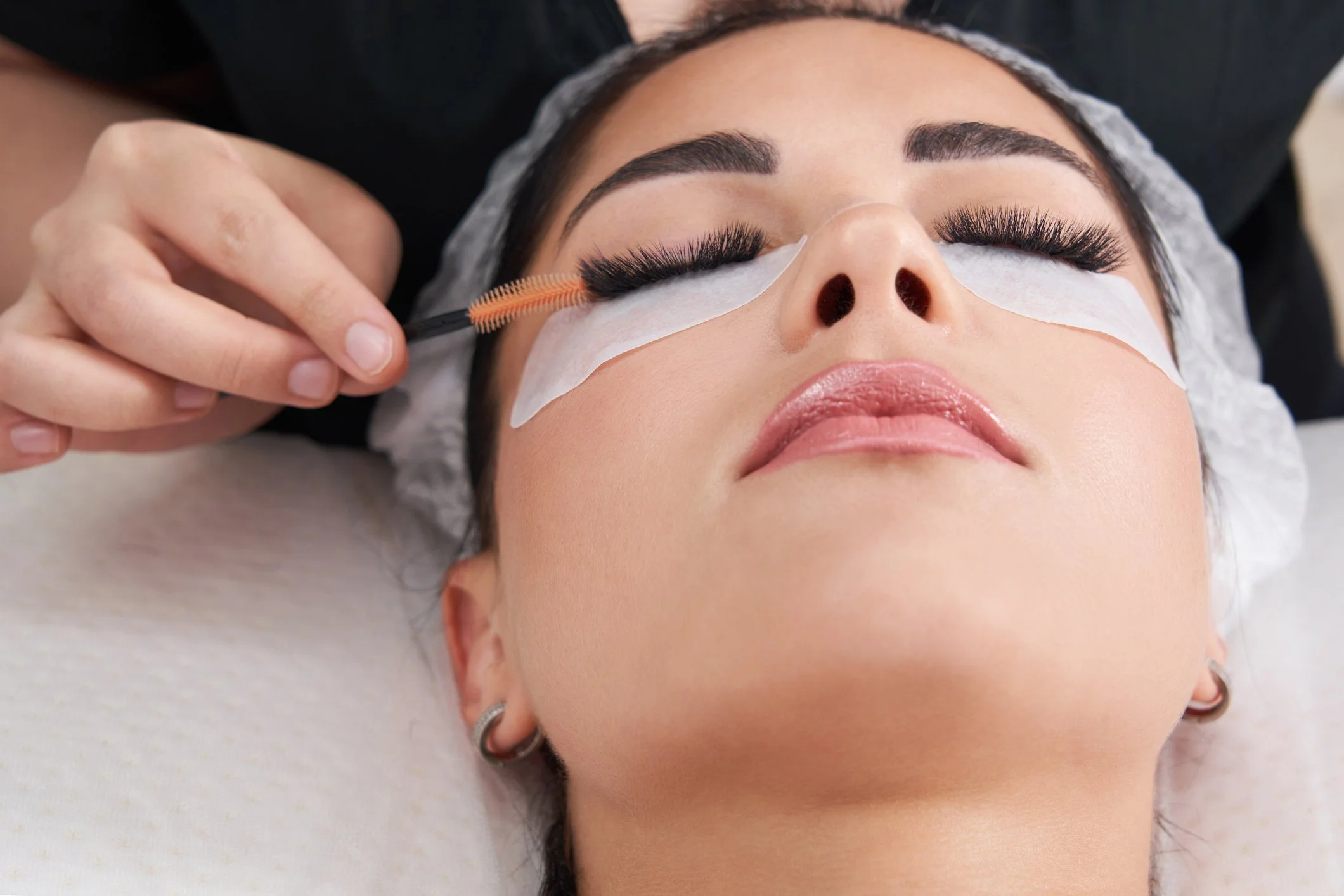 A woman lying on a bed with her eyes closed, having eyelash extensions applied, while a person with neatly trimmed nails uses a tool to attach the extensions.