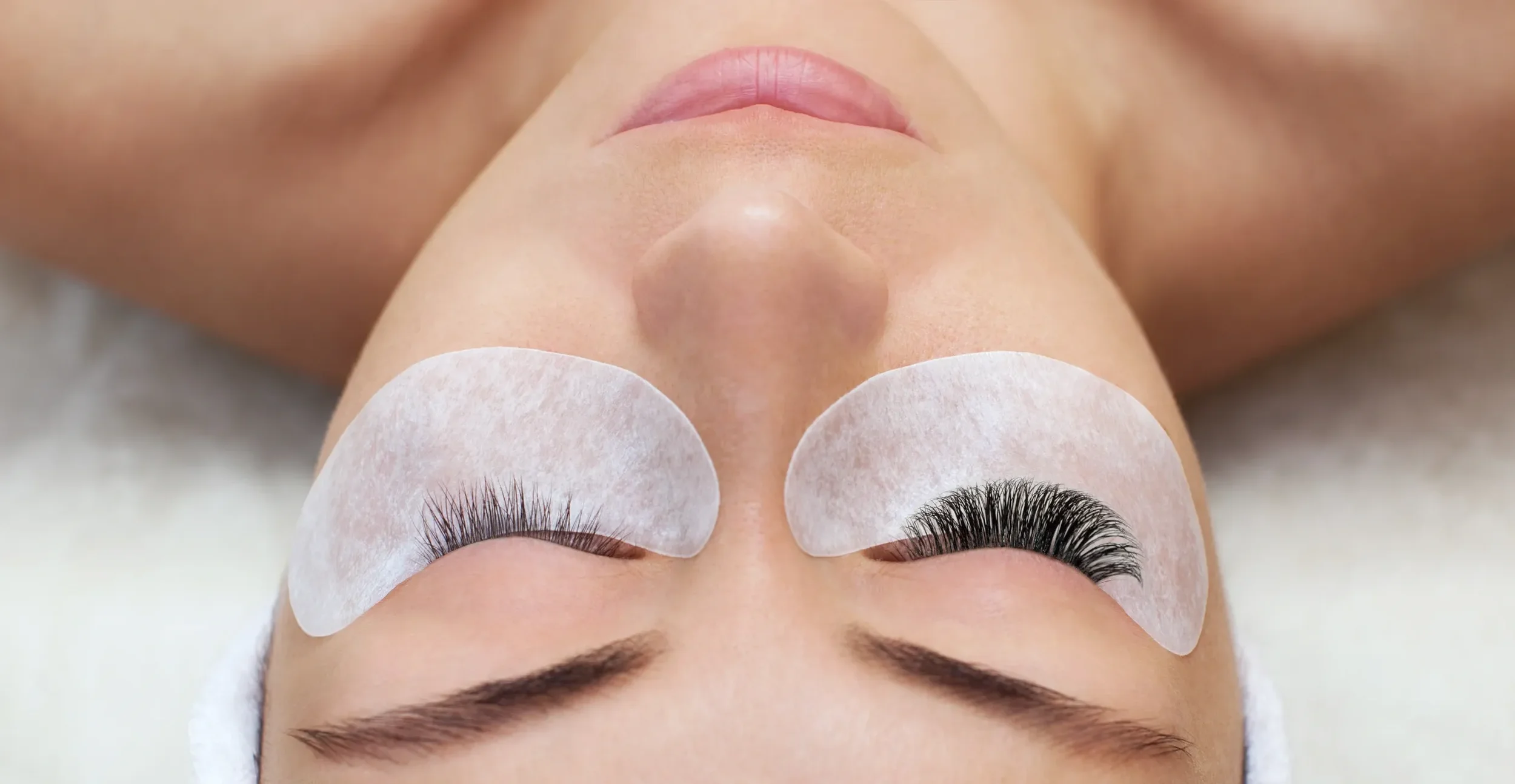 A woman lying down with eyes closed, receiving eyelash extensions while wearing eye patches and a facial mask.