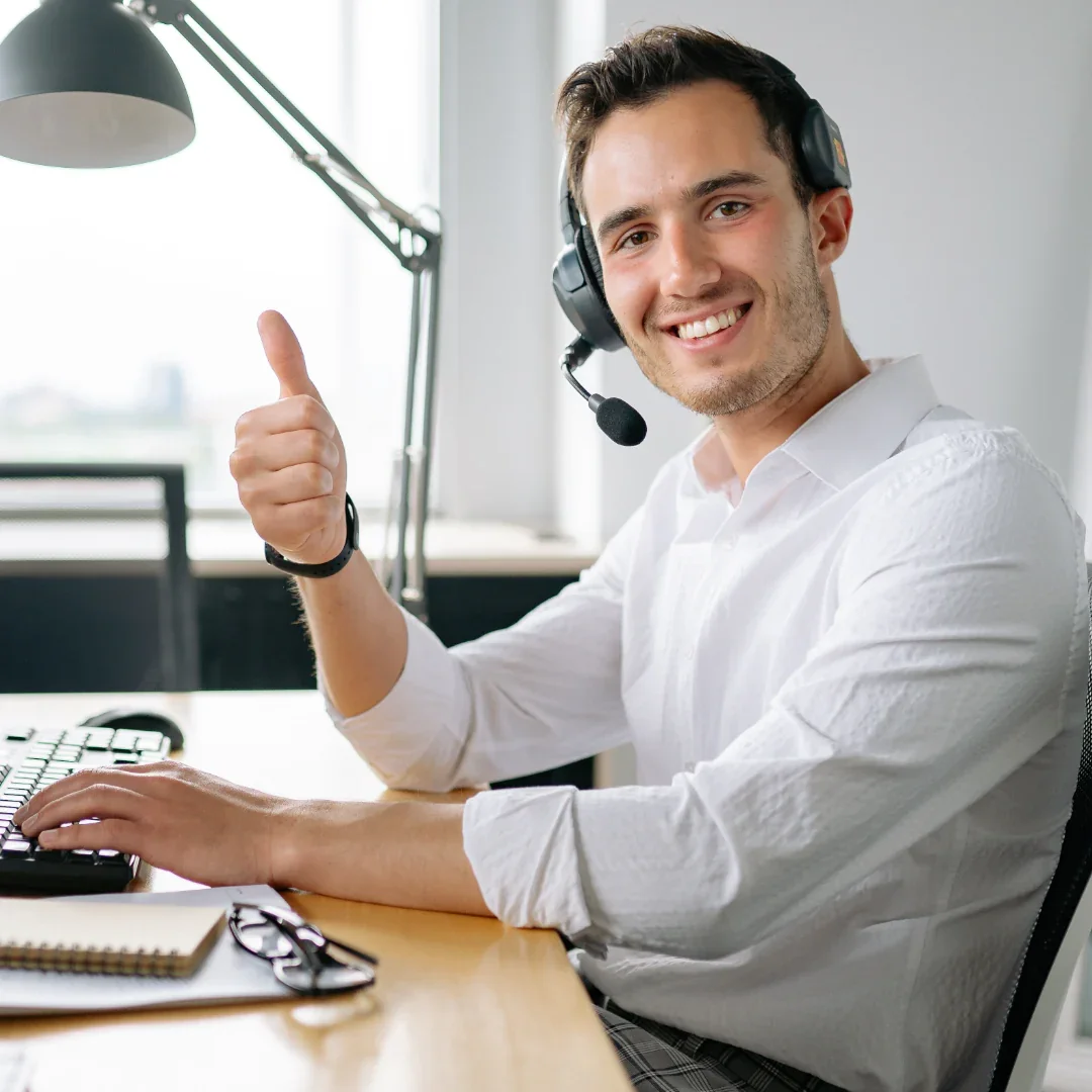 Giovane uomo sorridente con cuffie con microfono, seduto a scrivania in ufficio, dando pollice in su.