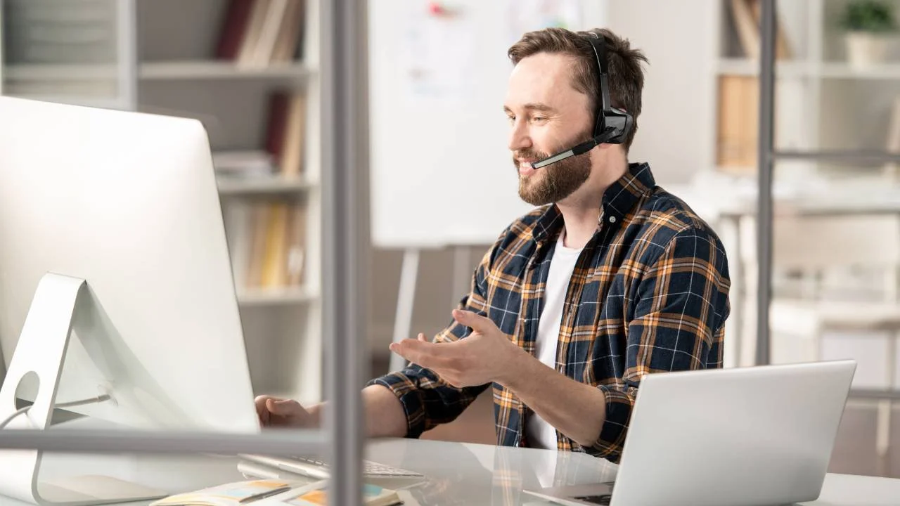 Uomo sorridente con cuffie e microfono, seduto a una scrivania mentre usa un computer