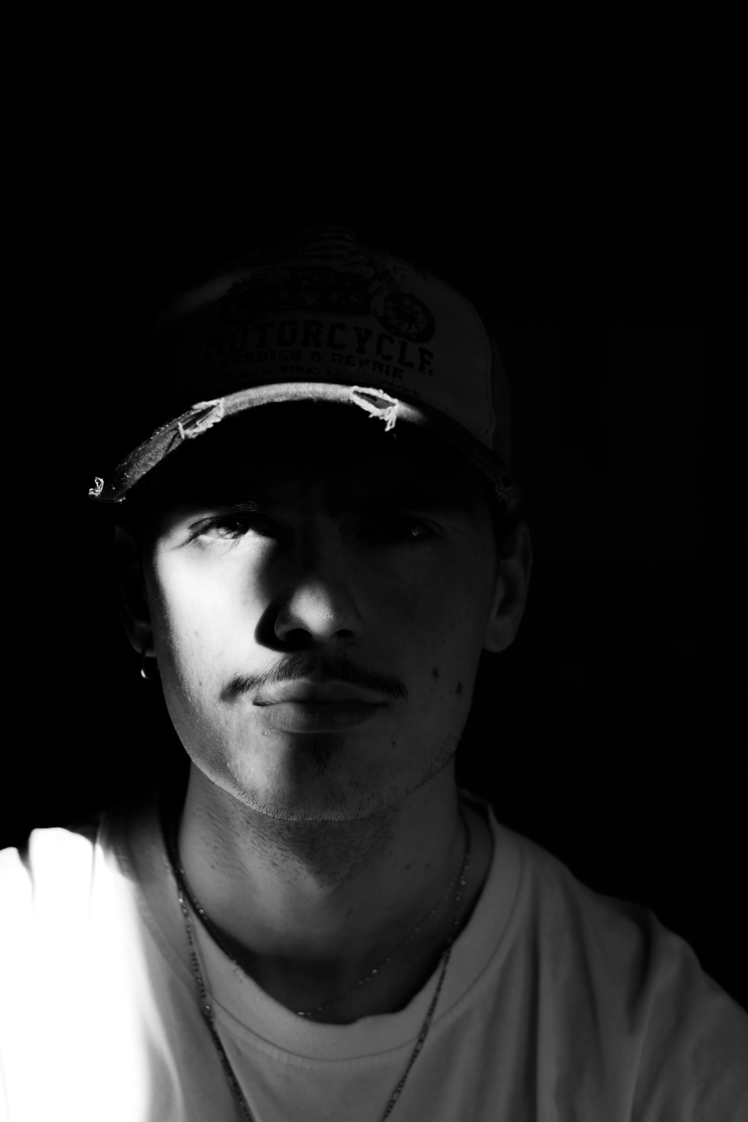 A black and white photo of a young man wearing a cap with a motorcycle logo. One side of his face is illuminated, creating a dramatic contrast with the dark background.